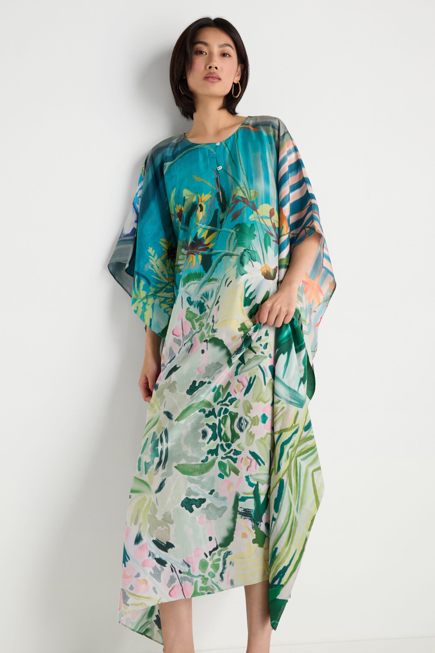 A person stands against a white wall wearing the Fern Silk Kaftan, featuring a loose, colorful botanical print with short sleeves and a relaxed, flowing fit.