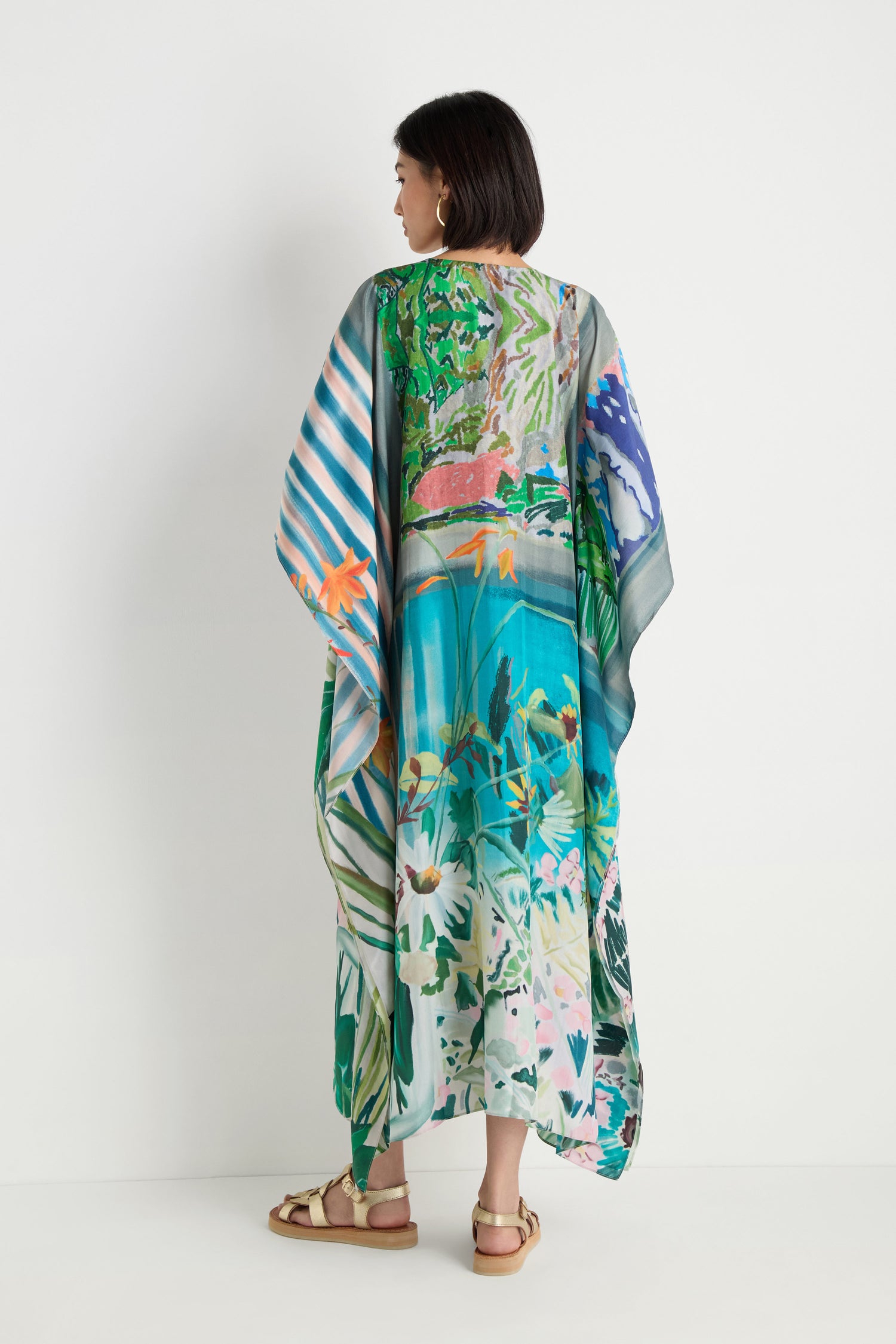 A woman stands against a white wall, shown from the back, wearing the Fern Silk Kaftan with a vibrant botanical print and flat sandals.