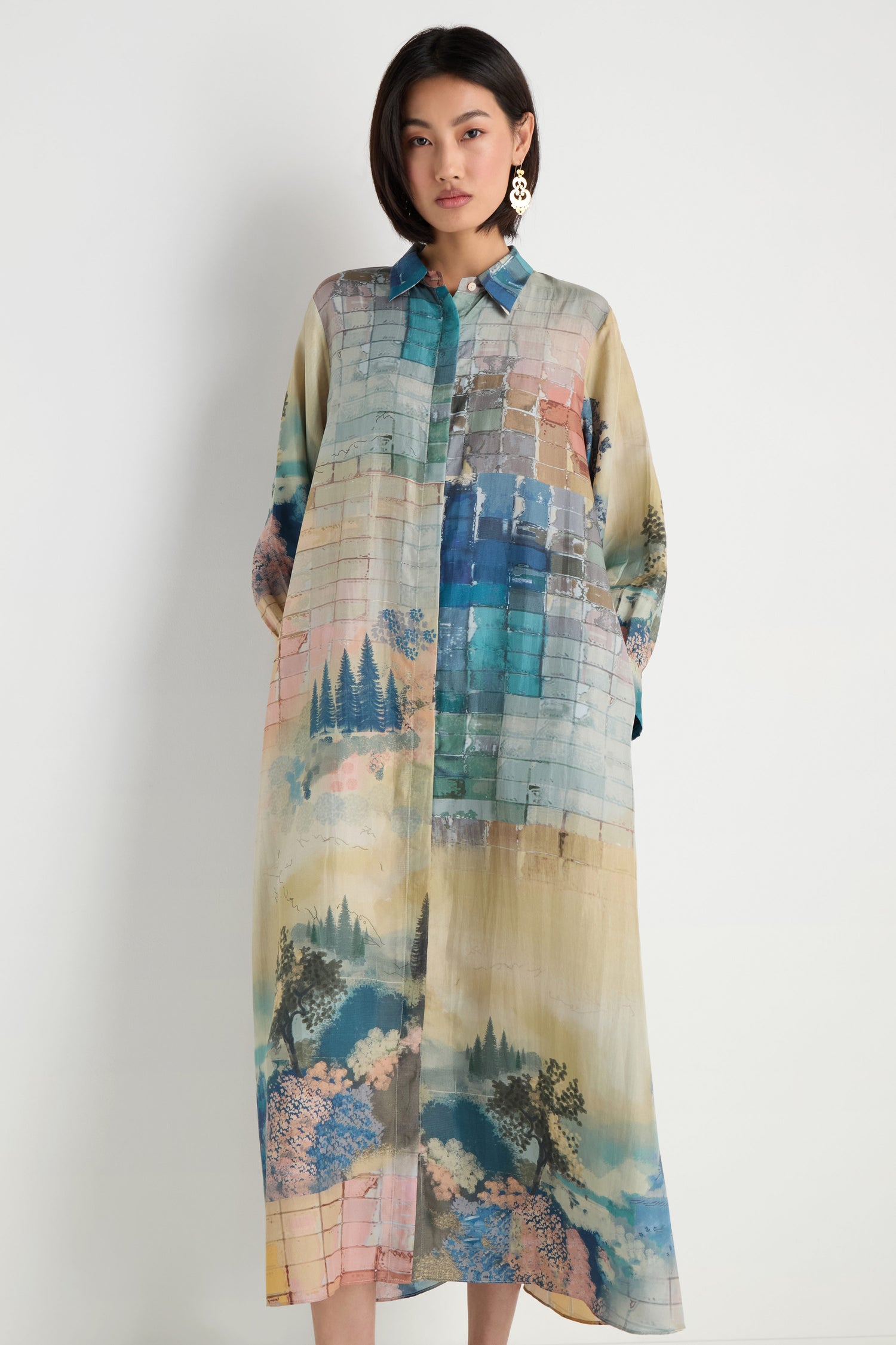 A woman wears the Horizon Silk Shirt Dress, a maxi-length, loose-fit piece with long sleeves, a collar, and scenic landscape and horizon prints in blue, green, and pastel tones.
