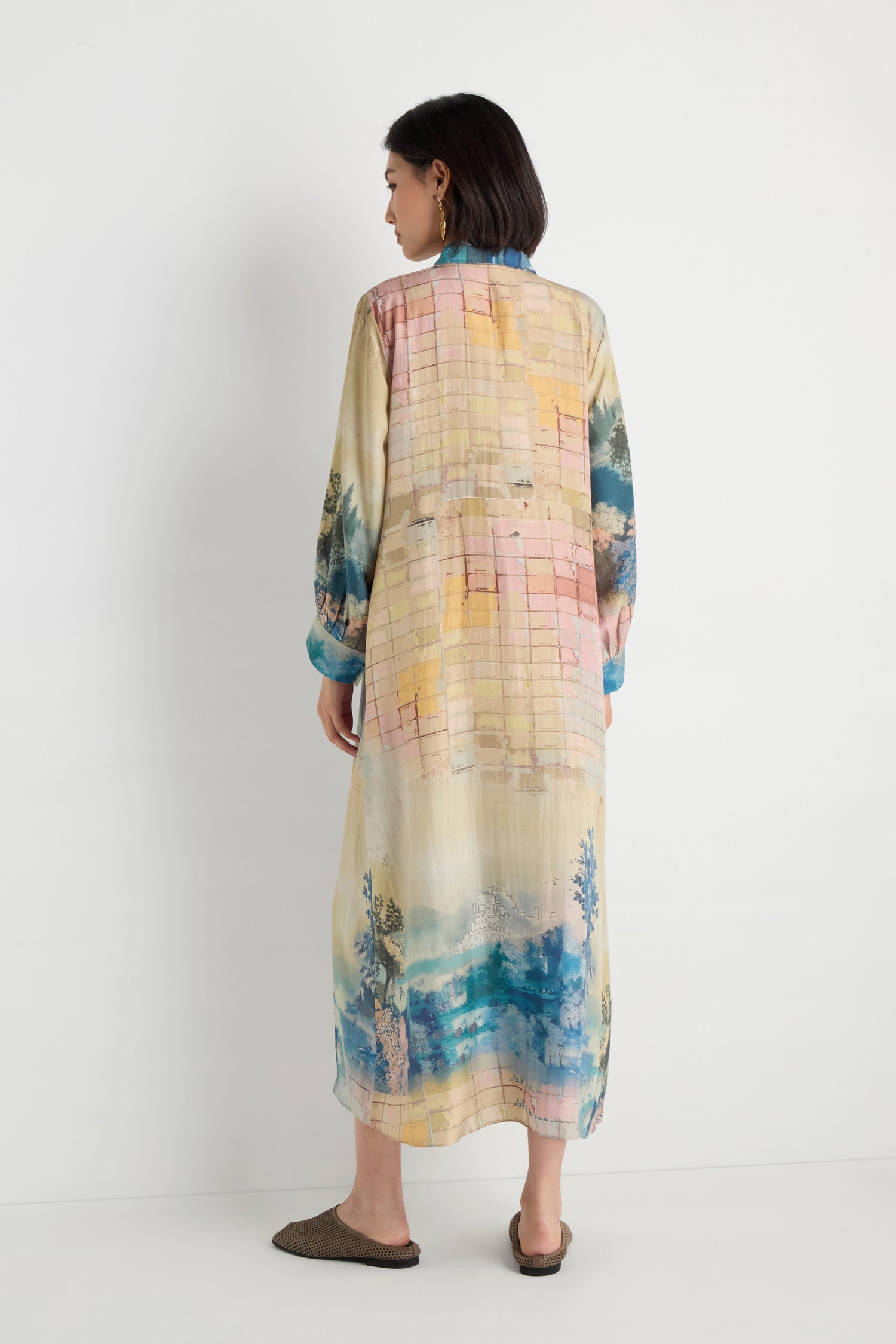 A woman stands facing away in the Horizon Silk Shirt Dress, a maxi-length piece featuring pastel abstract and nature-inspired patterns, paired with brown slip-on shoes against a plain white background.