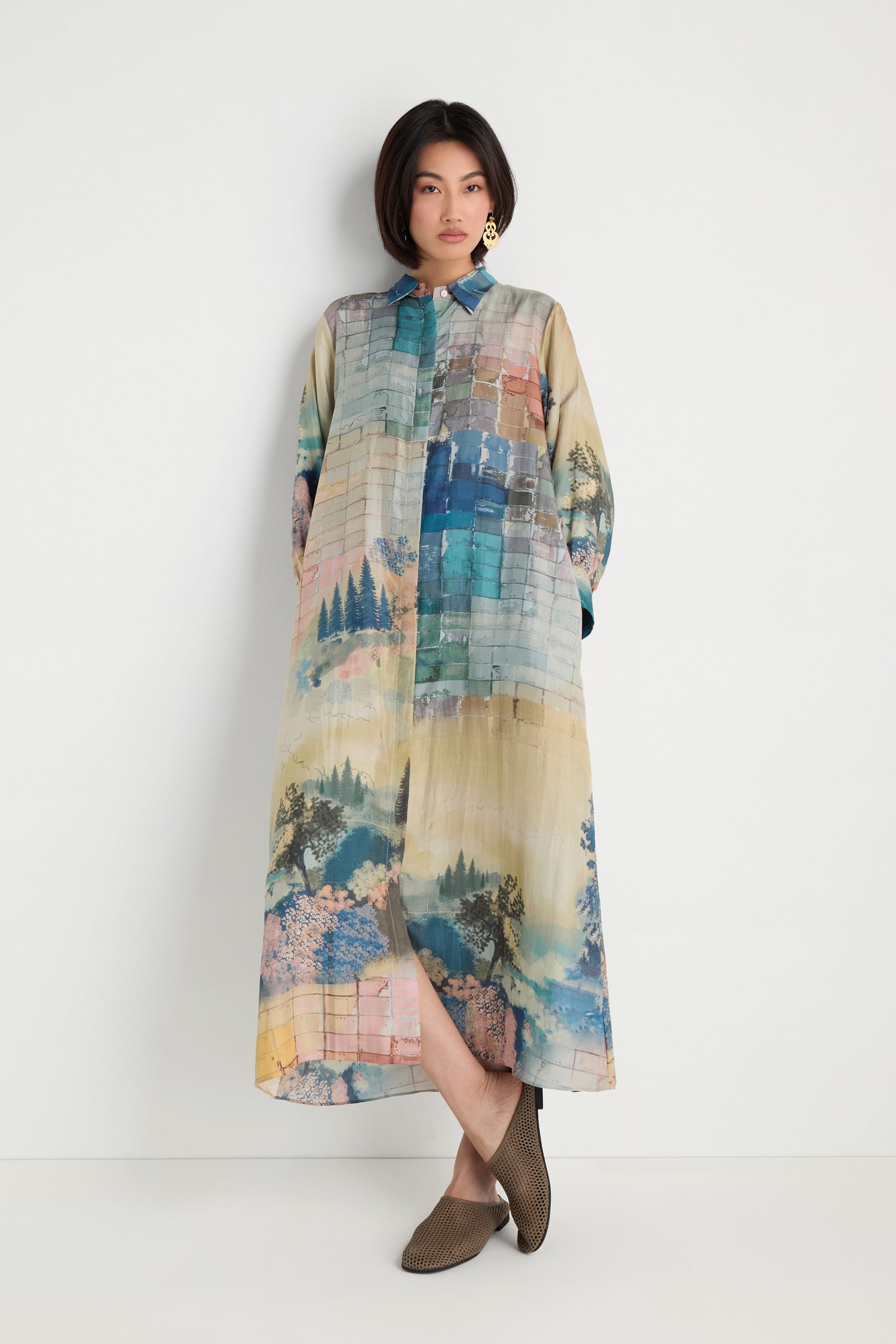 A person stands against a white wall wearing the Horizon Silk Shirt Dress, featuring a vibrant horizon print and maxi length, paired with brown shoes.