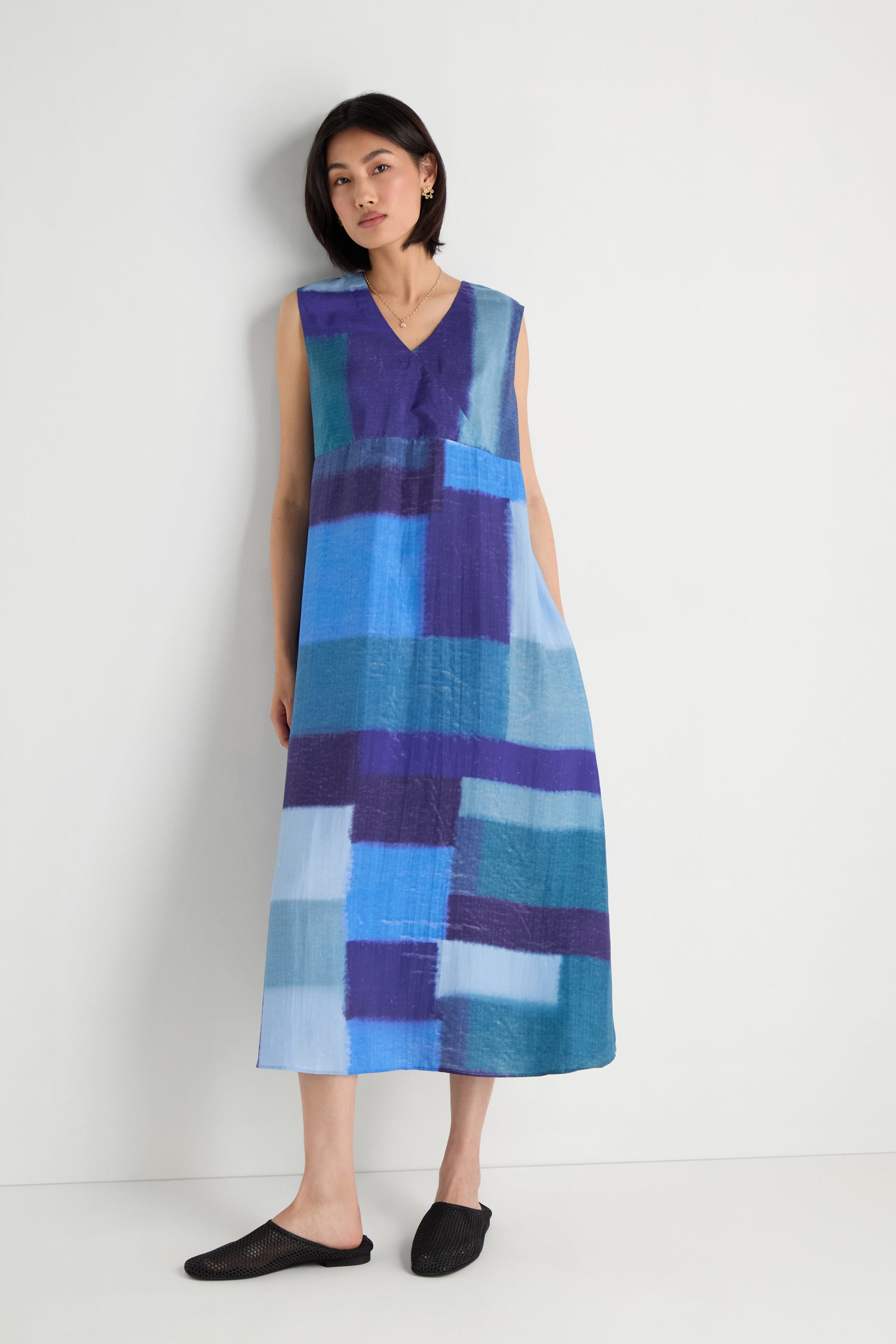 A woman stands against a plain white wall wearing the Blurred Blocks Swing Dress, a sleeveless, midi-length silk dress featuring a blue and purple blurred block print pattern.