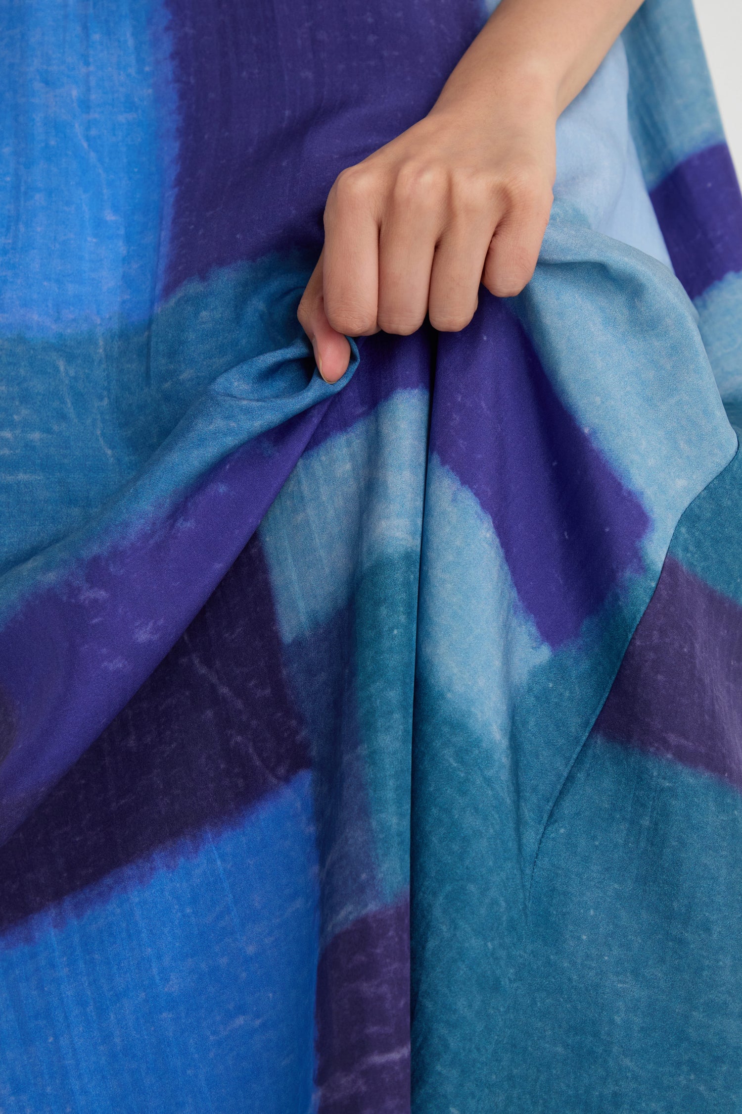 A hand holds a section of the Blurred Blocks Swing Dress, showcasing its blue and purple tie-dye silk, soft texture, and subtle block print details.
