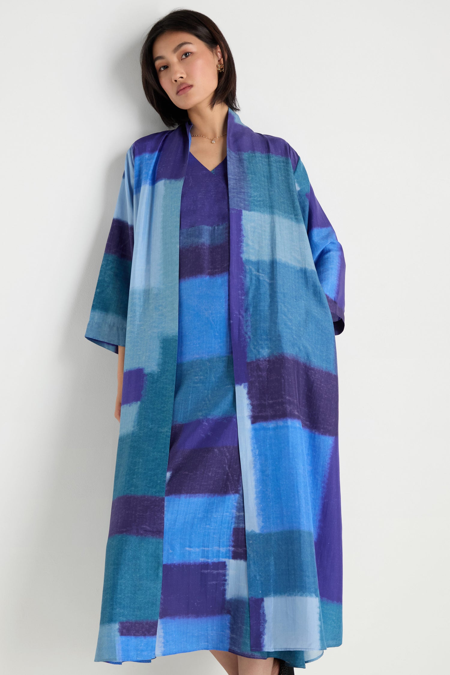 A woman stands against a white wall wearing the Blurred Blocks Easy Jacket—a long, open-front layer featuring a blue and purple color-block pattern, perfect for lightweight layering.
