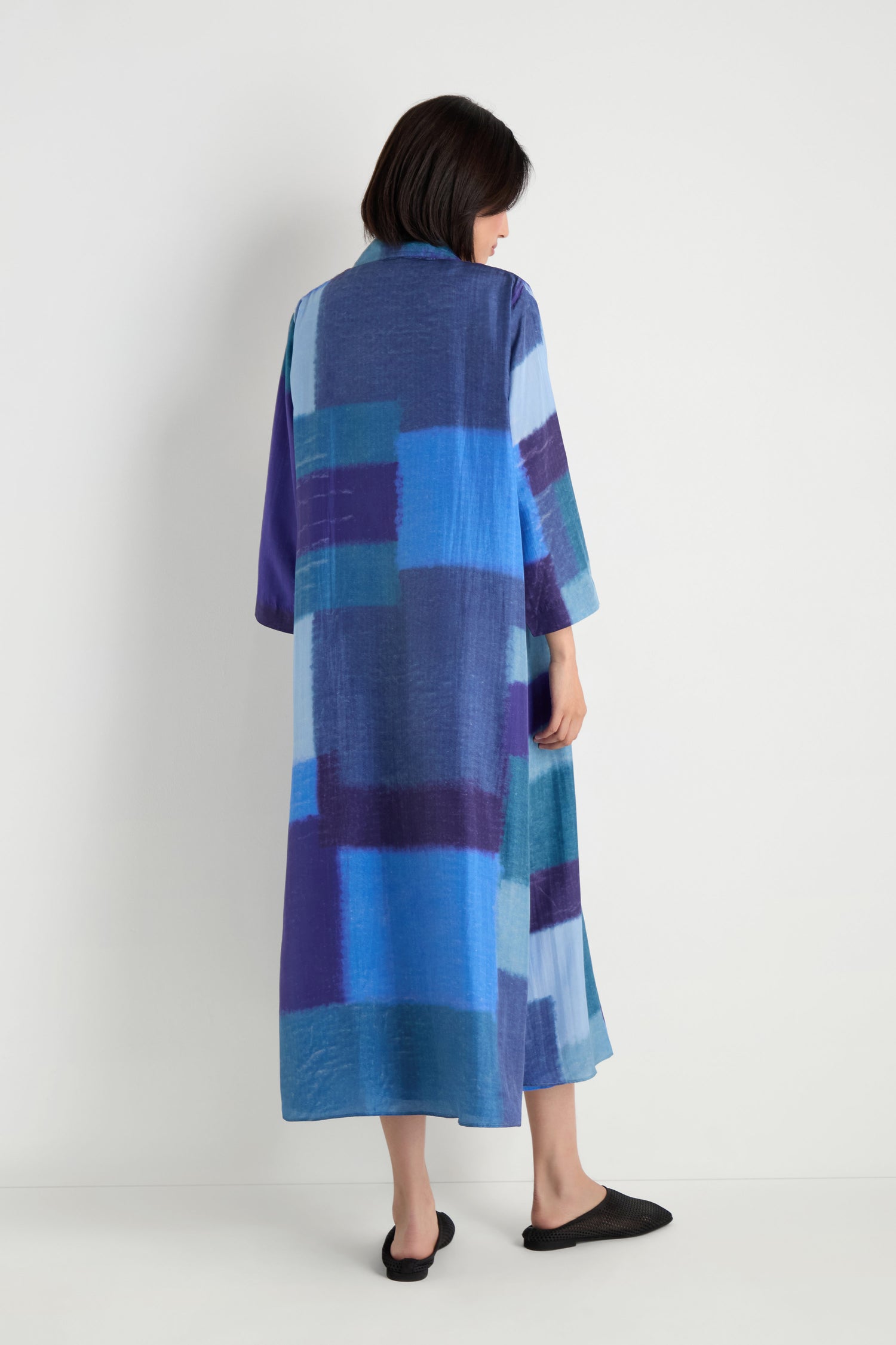 A person with short dark hair wears the Blurred Blocks Easy Jacket over a long, loose blue and purple geometric dress, paired with black slip-on shoes, standing facing away.