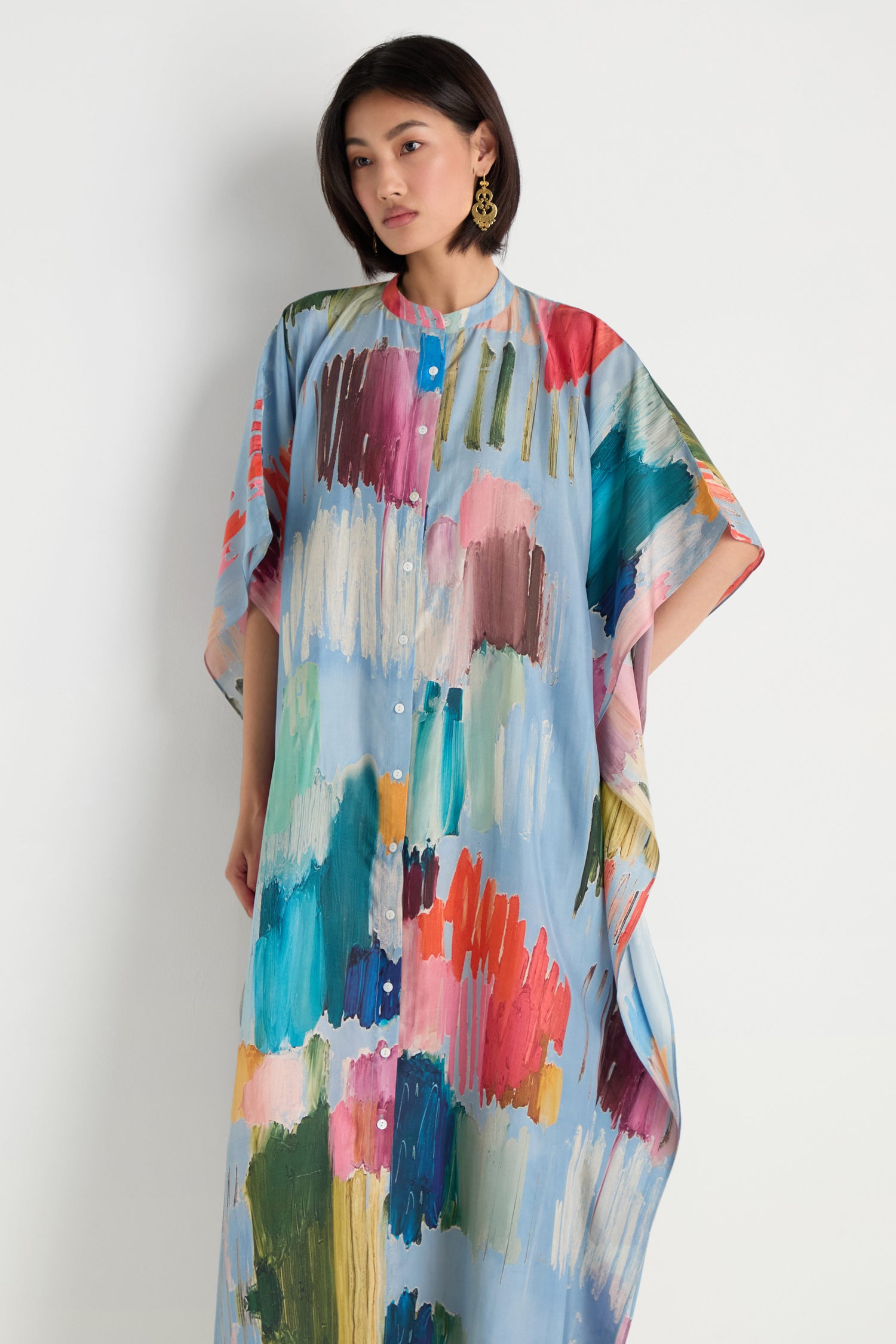A woman models the Breezy Strokes Silk Shirt Dress, featuring a loose fit and a vibrant abstract brushstroke print, standing against a plain white background.
