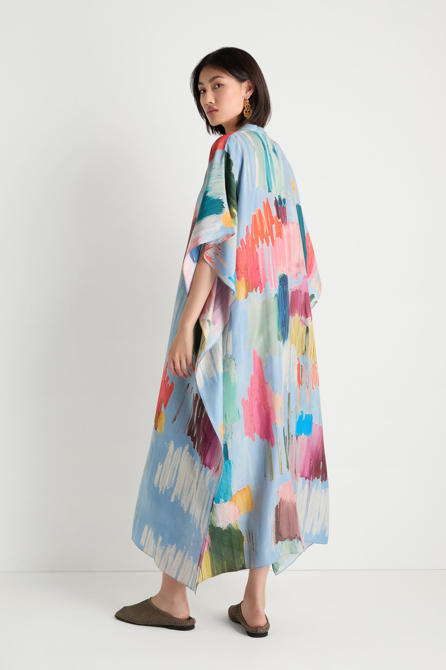 A woman stands against a white wall, looking over her shoulder, wearing the Breezy Strokes Silk Shirt Dress in colorful hues with brown slip-on shoes.
