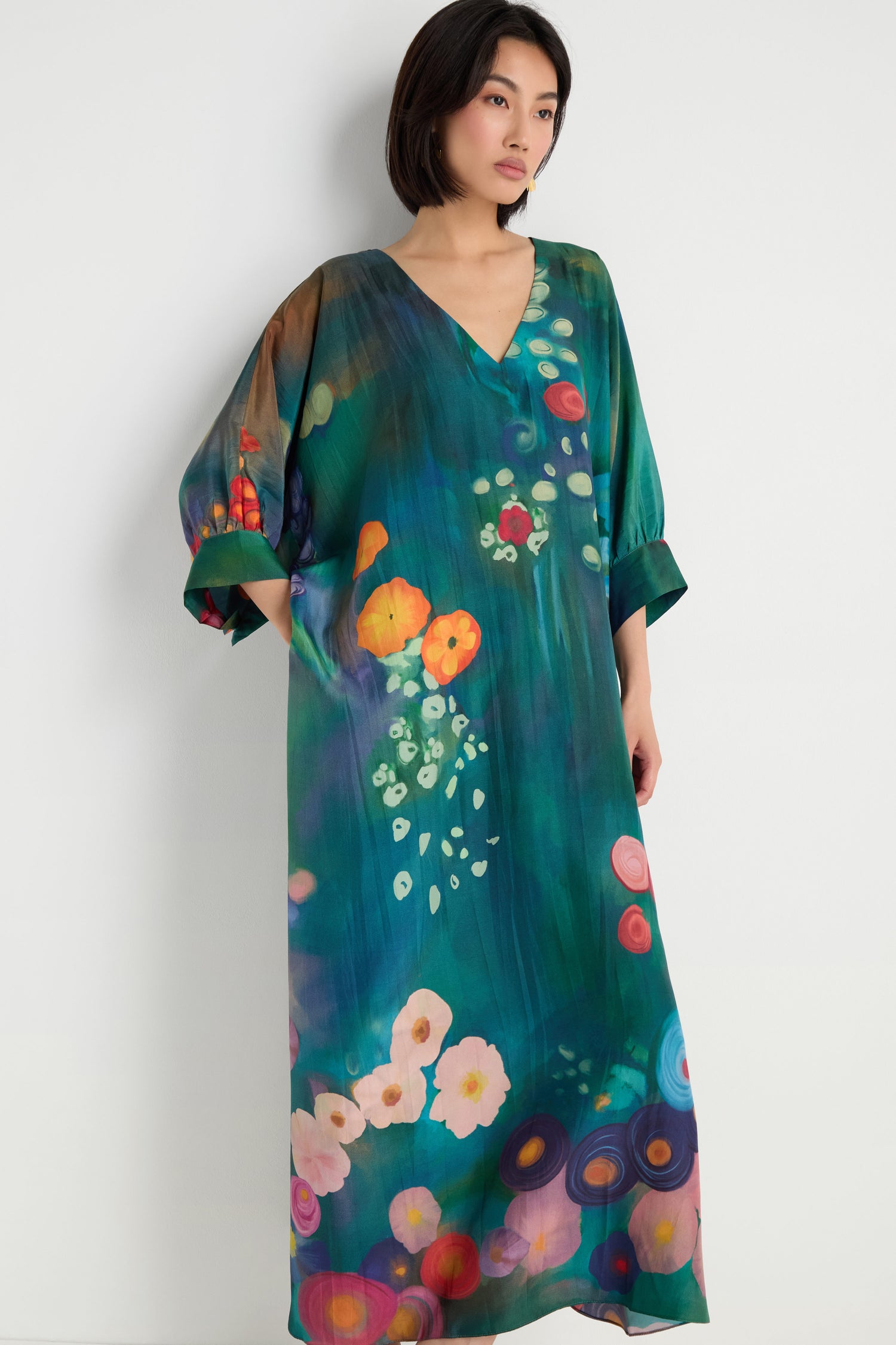 A woman stands against a plain wall wearing the Soft Floral Silk V-Neck Dress, featuring a loose fit and an abstract floral print in vibrant green, blue, red, and orange tones.