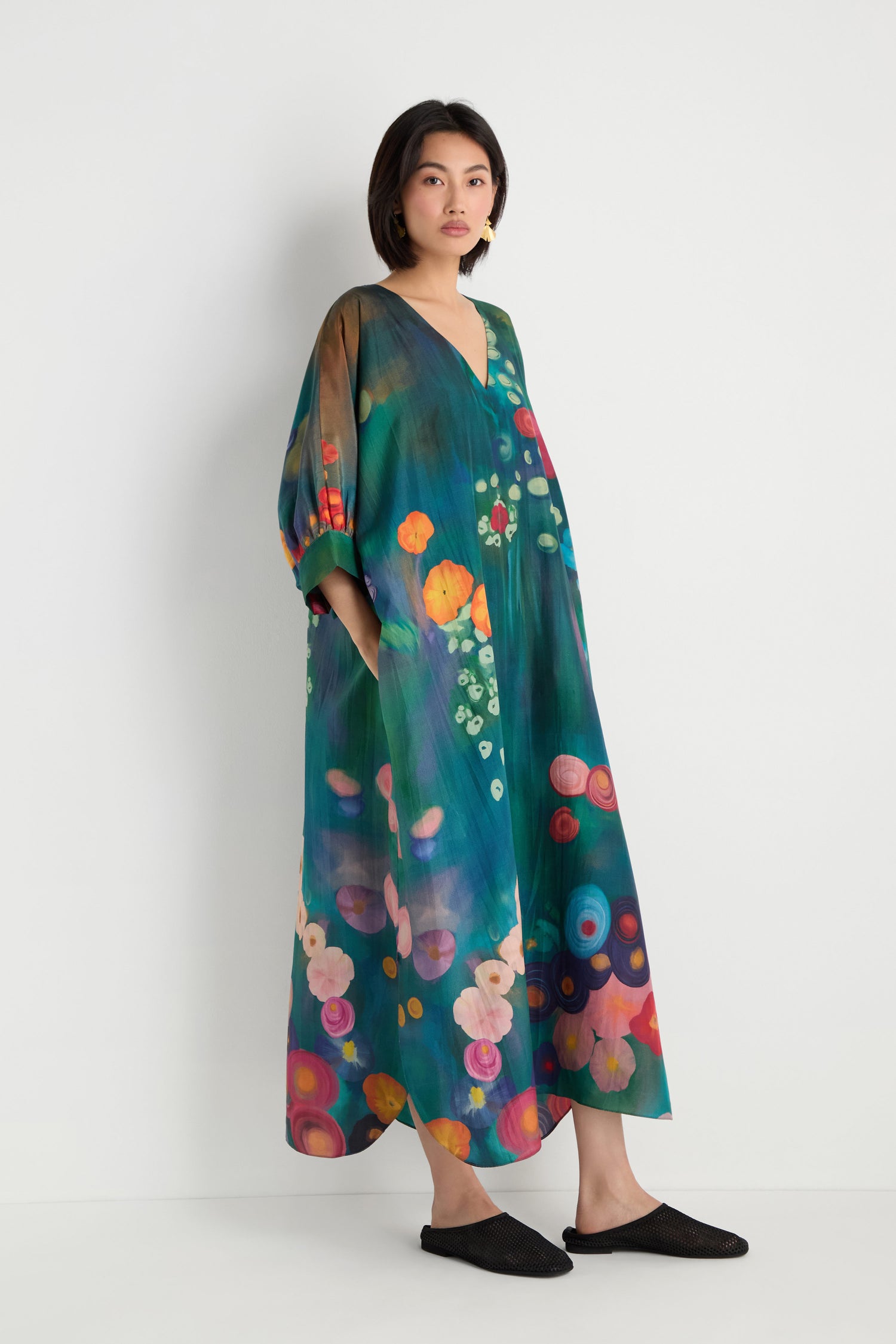 A woman stands against a white wall wearing the Soft Floral Silk V-Neck Dress, an ankle-length, loose silk dress with an abstract floral print, paired with black flat shoes.