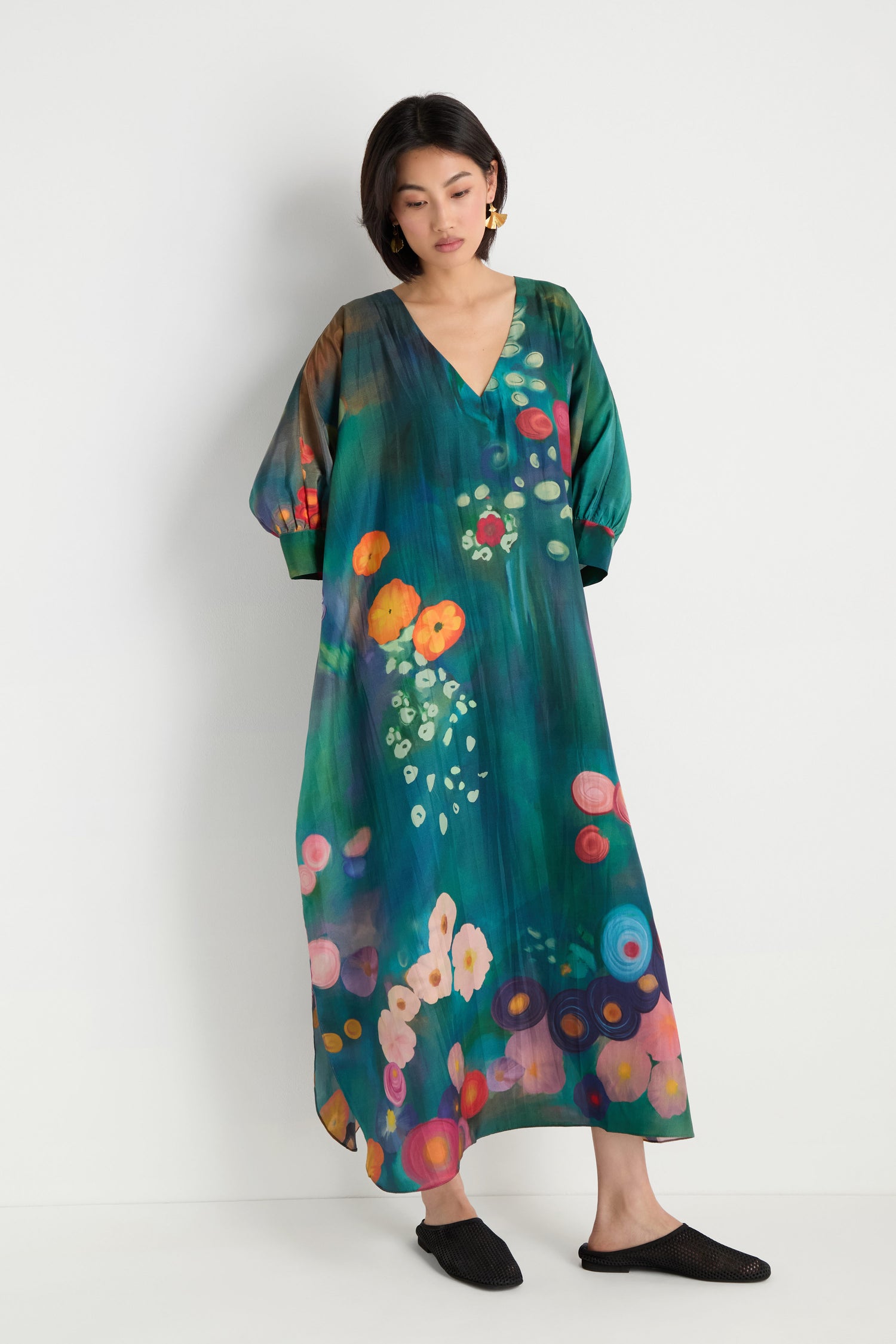 A woman stands against a white wall wearing the Soft Floral Silk V-Neck Dress, featuring a long, loose fit with an abstract floral print, paired with black flat shoes.
