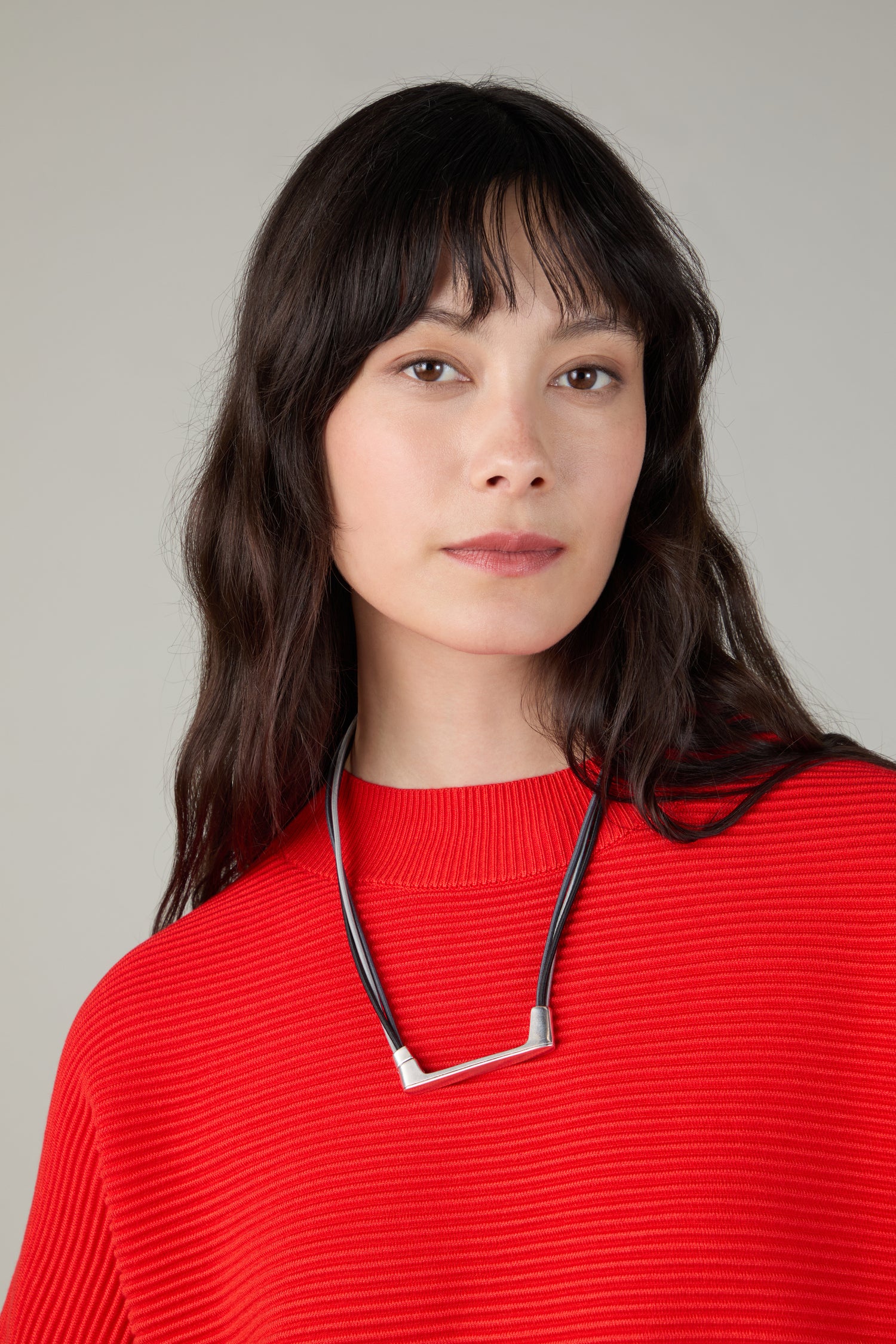A woman with long dark hair and bangs wears a bright red ribbed sweater and the Versatile Triple Cord Bracelet, standing against a neutral background.
