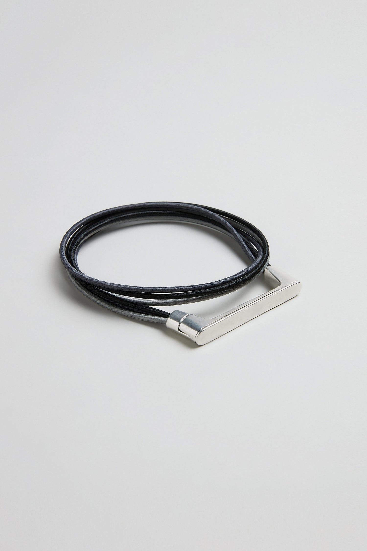 A Versatile Triple Cord Bracelet, crafted from black leather with a sleek silver rectangular bar, is coiled on a plain light background, reflecting the elegant style of Christina Brampti.