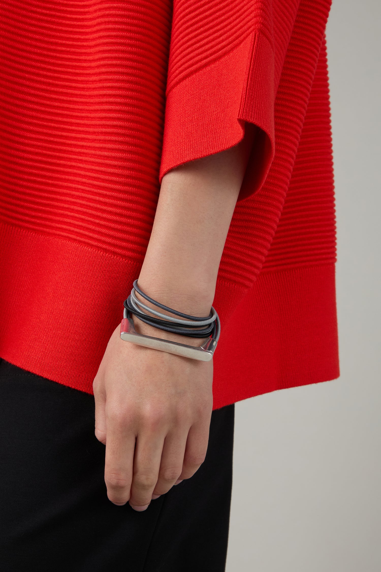 A person wears a red ribbed top, black pants, and several metal and leather bracelets, including the Versatile Triple Cord Bracelet on their wrist.