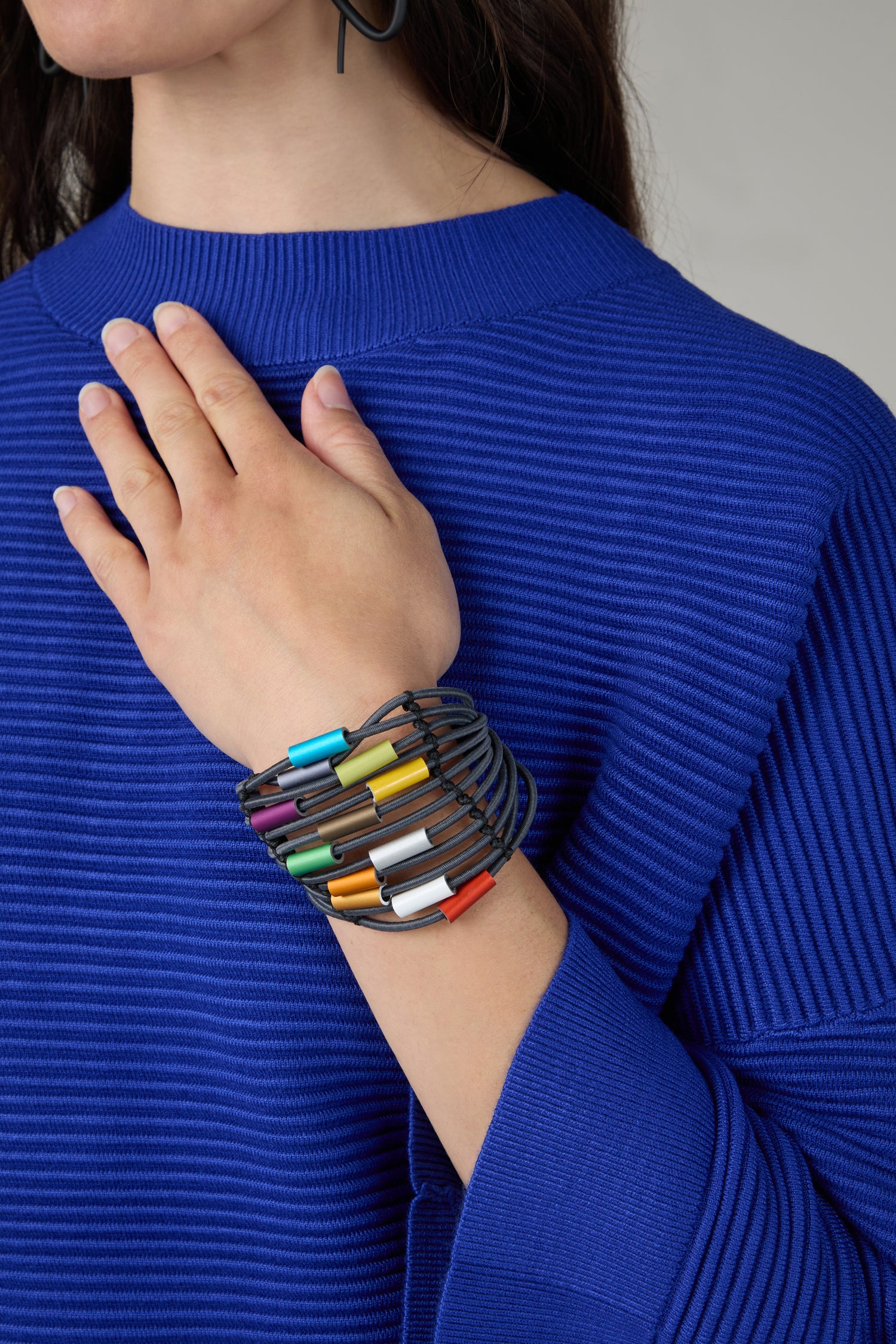 A person in a textured blue sweater touches their shoulder, showcasing the Colour Pop Cylinders Bracelet—a contemporary multi-strand accessory adorned with vibrant geometric beads.