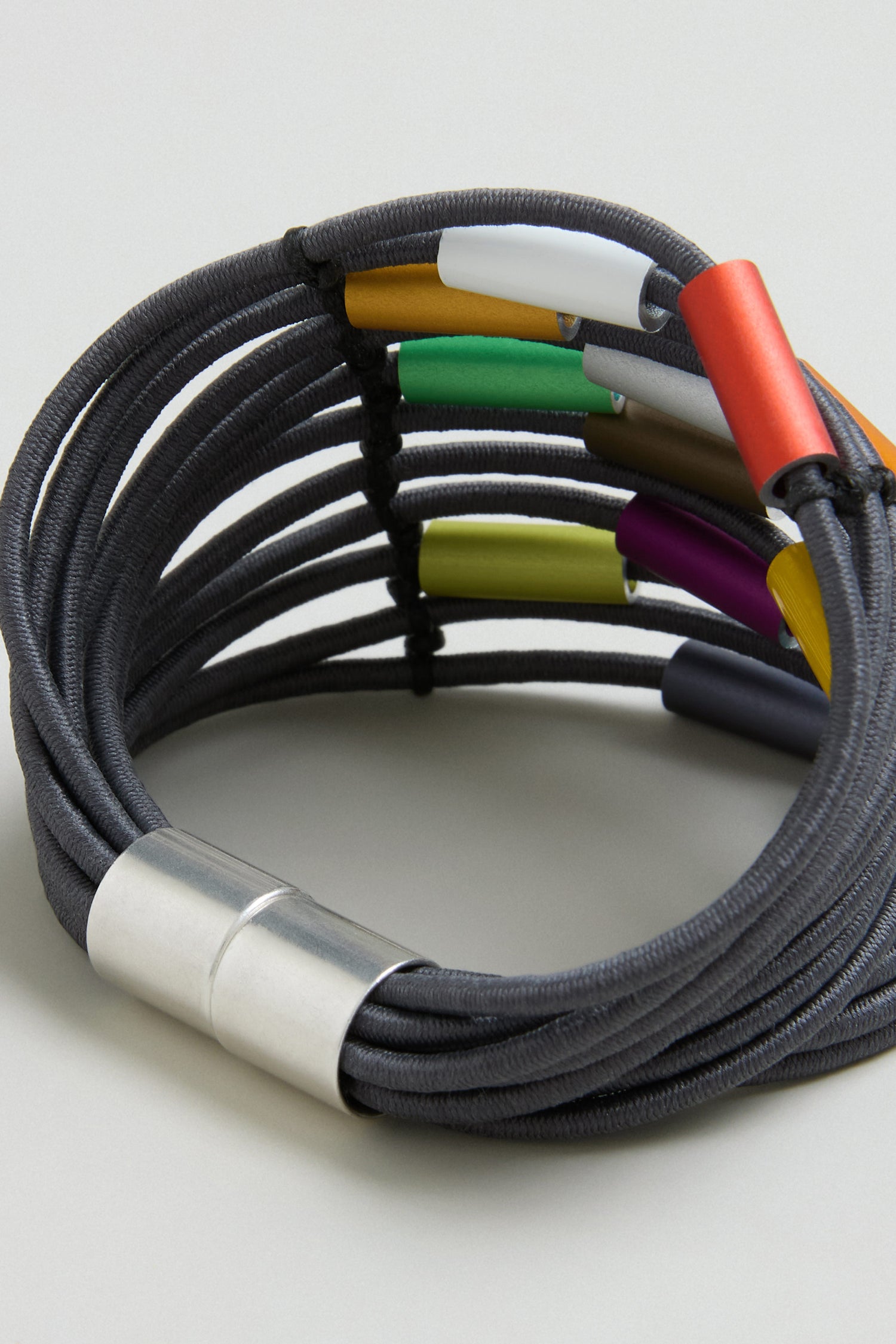 The Colour Pop Cylinders Bracelet by Christina Brampti features multi-strand black cords, colorful metal tube accents, and a silver magnetic clasp, displayed on a light surface for a modern look.