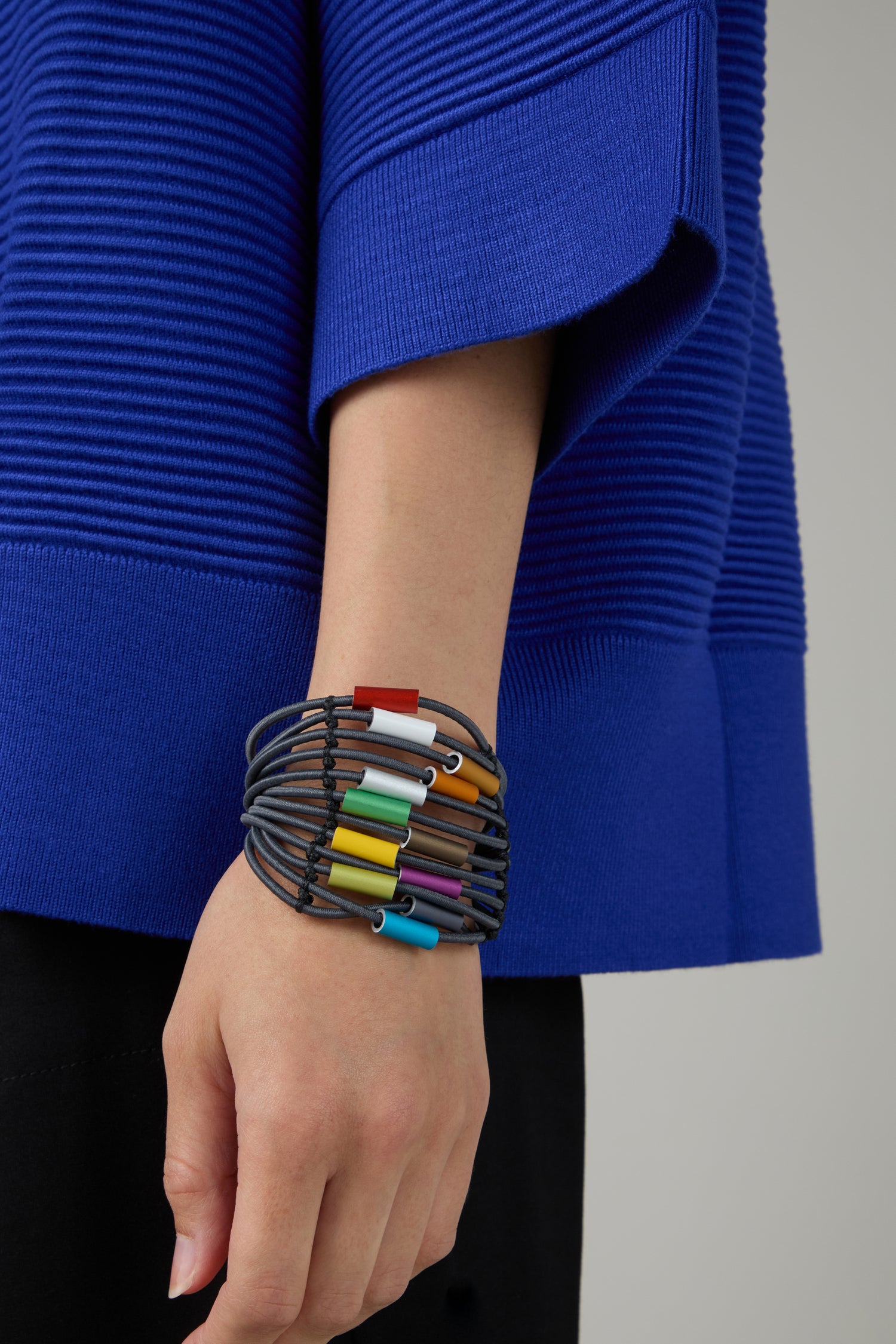 A person in a blue textured top wears the Colour Pop Cylinders Bracelet by Christina Brampti, featuring contemporary jewellery with vibrant multi-strand cylindrical beads on their wrist.