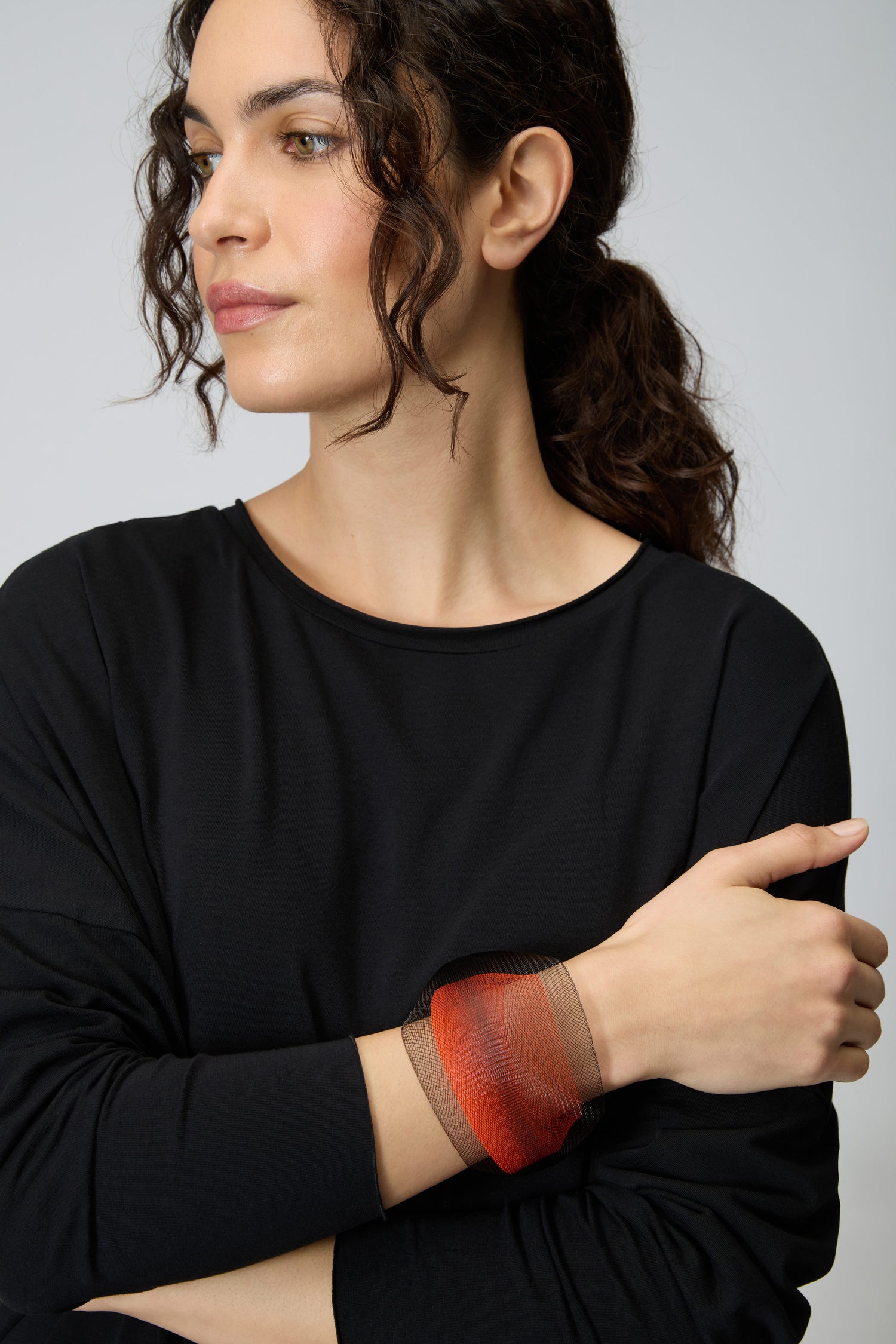 A woman with dark curly hair in a ponytail, wearing a black long-sleeve top and the Orange Statement Mesh Bracelet, stands against a plain background with arms crossed, channeling the bold style of Christina Brampti accessories.