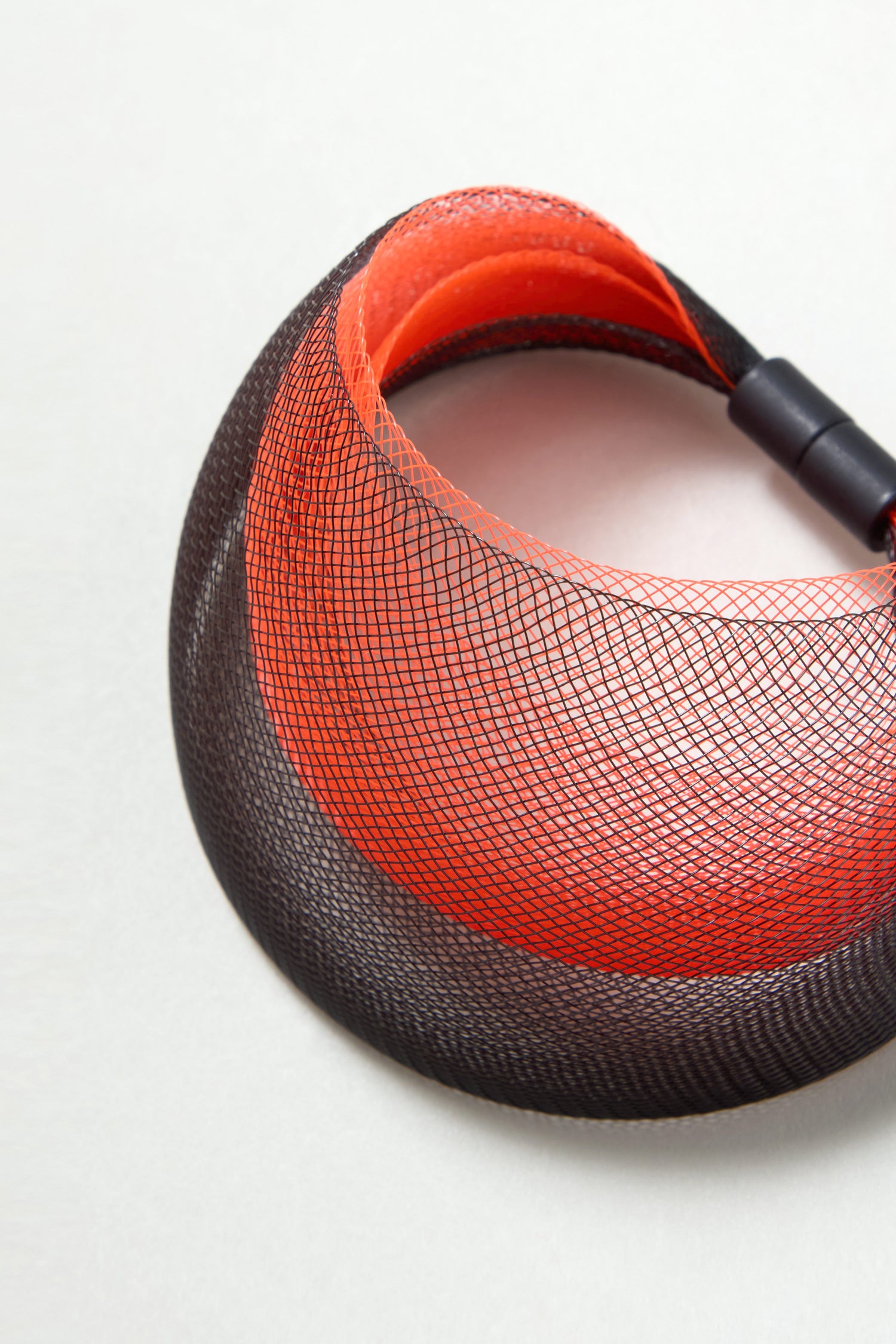 The Orange Statement Mesh Bracelet features a sculptural black and orange mesh design by Christina Brampti, coiled on a light surface and secured with a plastic fastener.