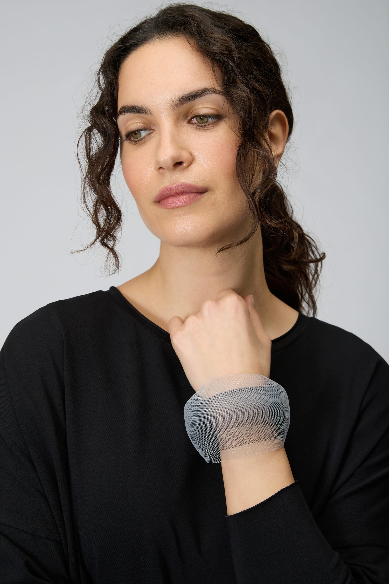 A woman with dark curly hair wears a black top and the Silver Statement Mesh Bracelet on her wrist, looking slightly down and to the side against a plain background.