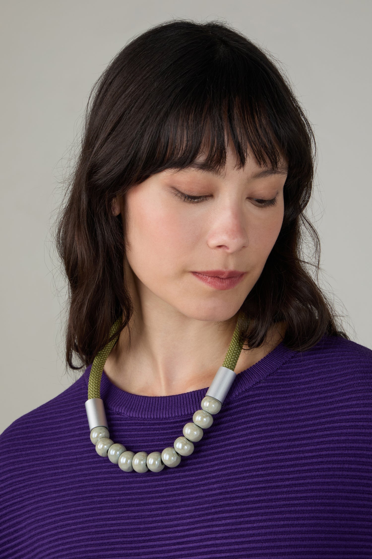 A woman with shoulder-length dark hair, wearing a purple ribbed top and the Olive Ceramic Milkyway Necklace, looks down with a neutral expression.