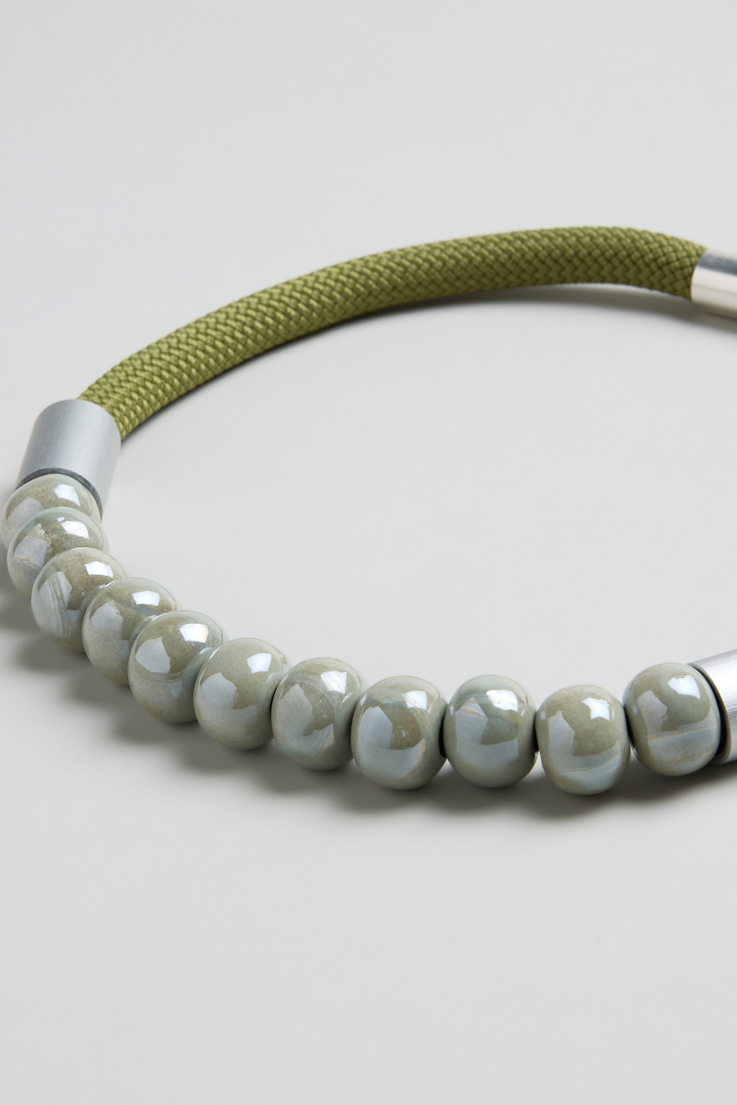 The Olive Ceramic Milkyway Necklace features glossy pale green beads on an olive green woven cord with silver metal end caps—a handcrafted piece that offers subtle, elegant style.