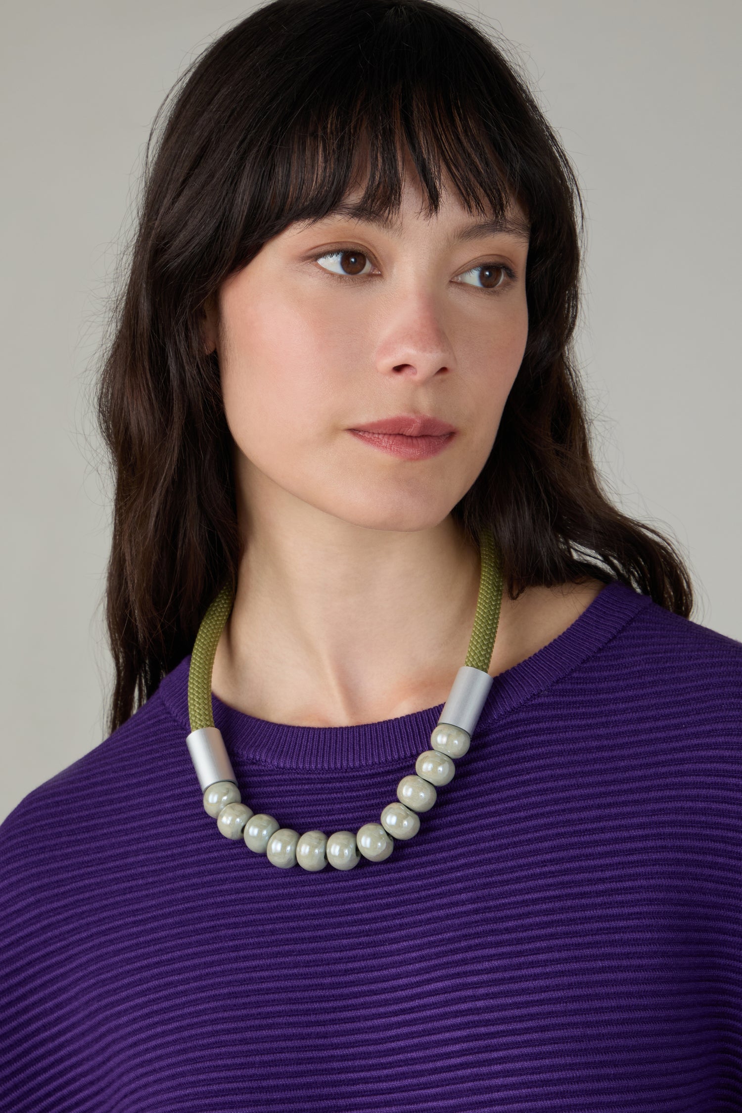 A woman with long dark hair, wearing a purple ribbed top and the Olive Ceramic Milkyway Necklace, featuring large metallic beads, looks slightly to the side against a neutral background.