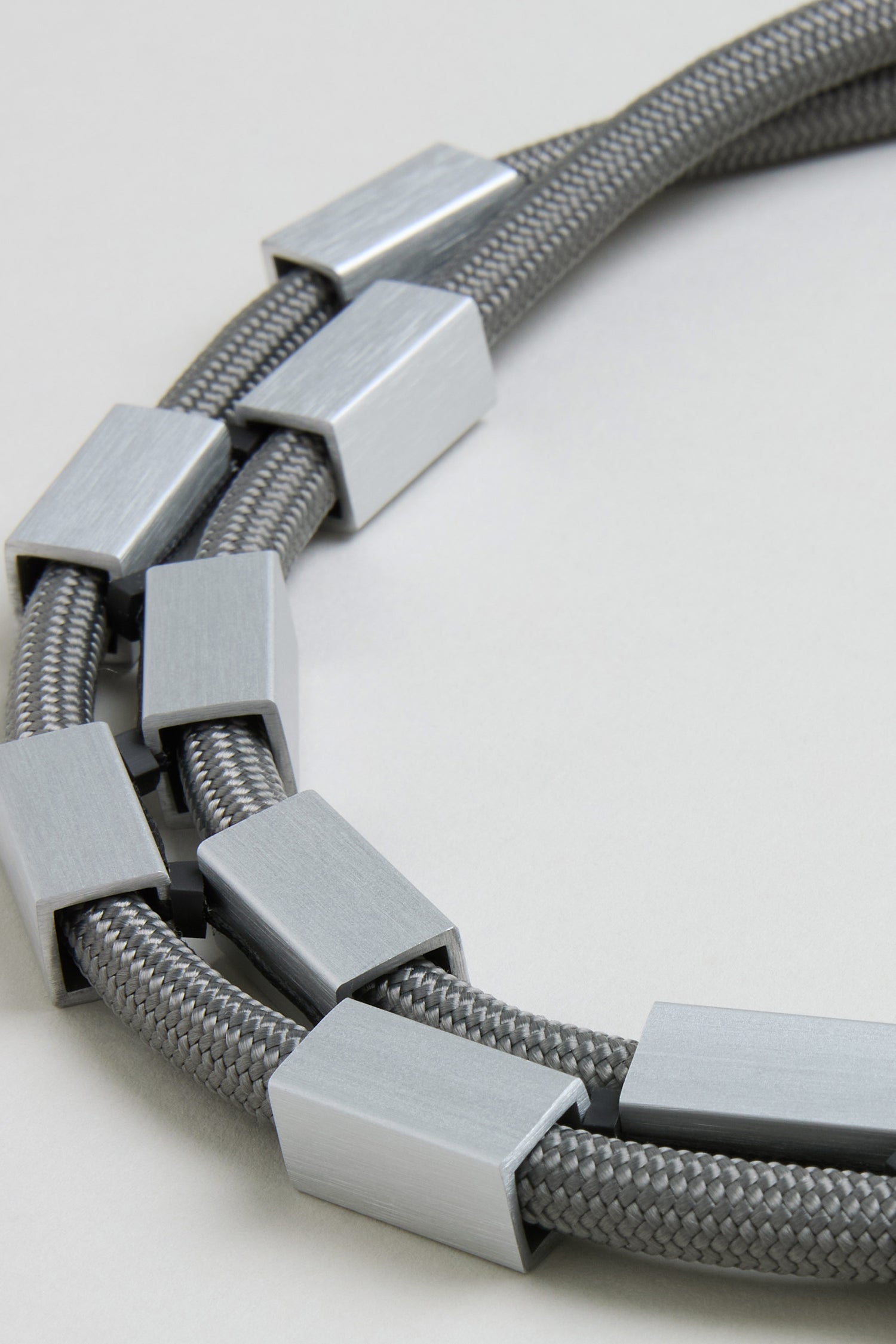 Close-up of the Short Aluminium Cube Cord Necklace, a gray handmade rope necklace with rectangular silver aluminum beads spaced along its length, shown on a light neutral background.