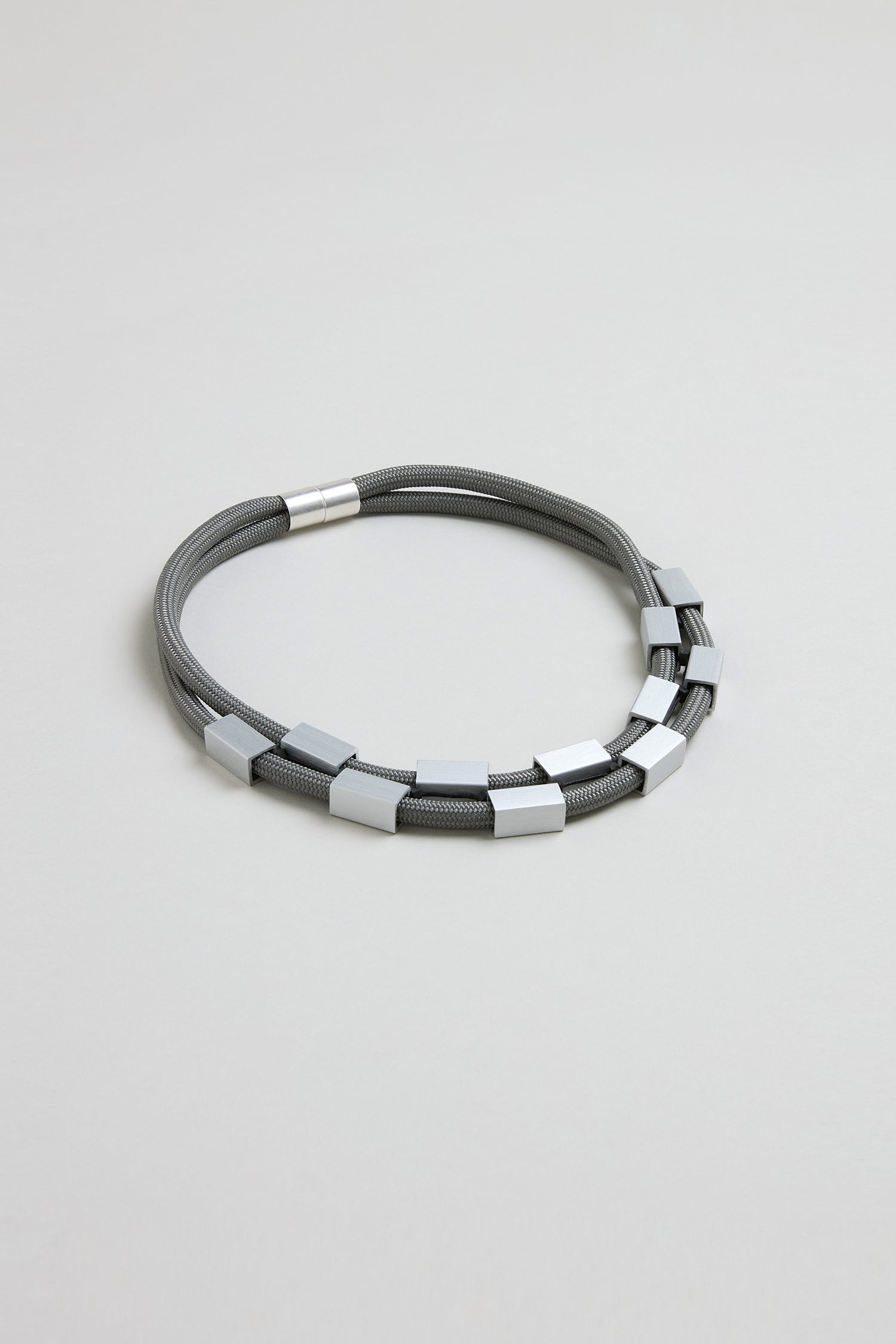 The Short Aluminium Cube Cord Necklace features gray multi-strand cords with evenly spaced aluminum cube beads and a cylindrical clasp, shown on a light gray background.