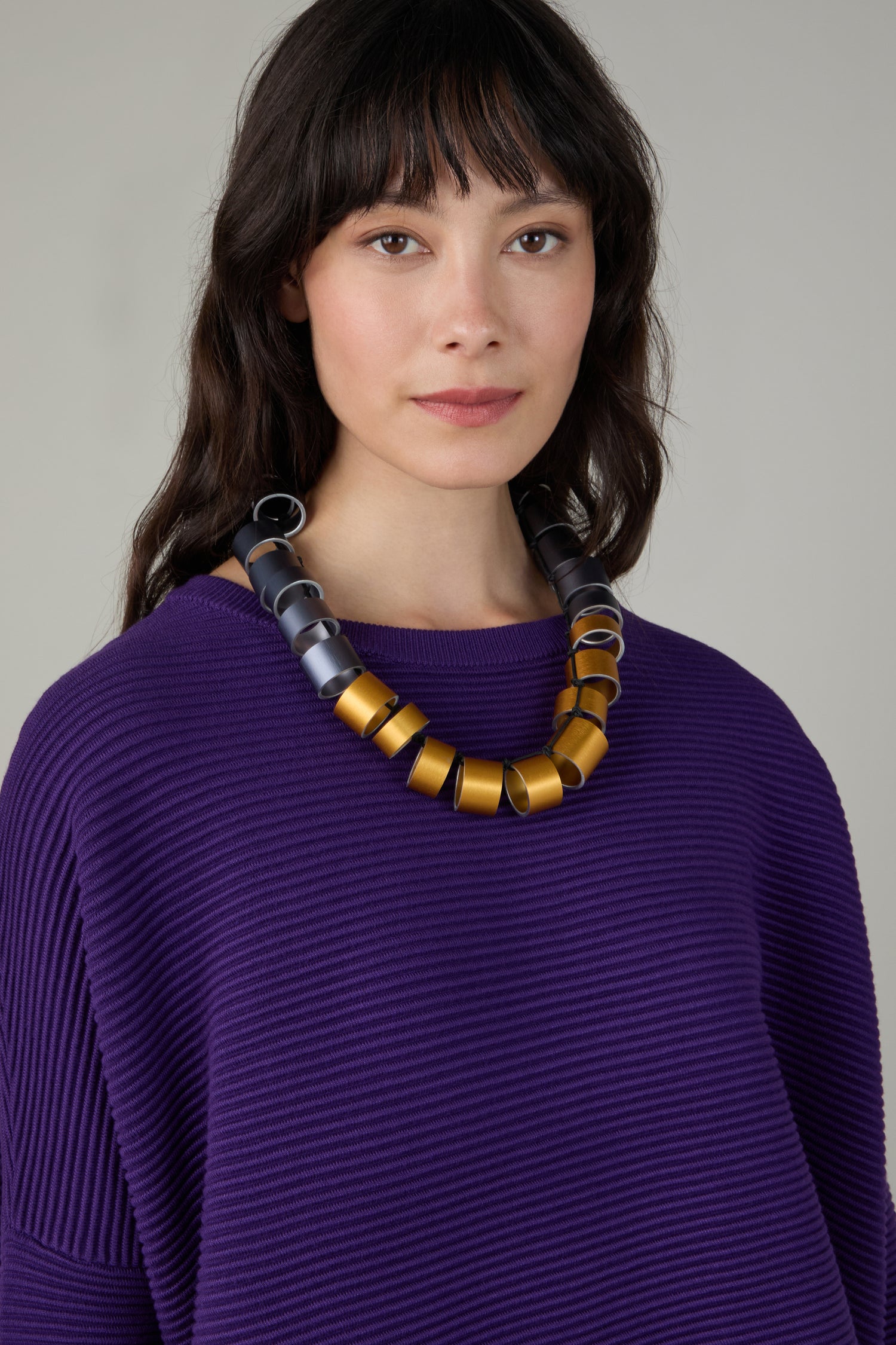 A woman with long dark hair wears a ribbed purple top and the Geometric Cylinders Necklace featuring bold gold and gray links, standing against a plain light background.