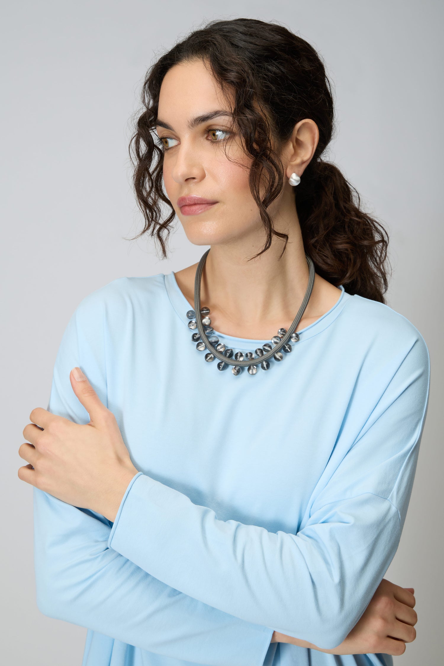 A woman with curly dark hair, in a light blue top and pearl earrings, stands with arms crossed, looking to the side and wearing the Smoke Atmosphere Necklace.