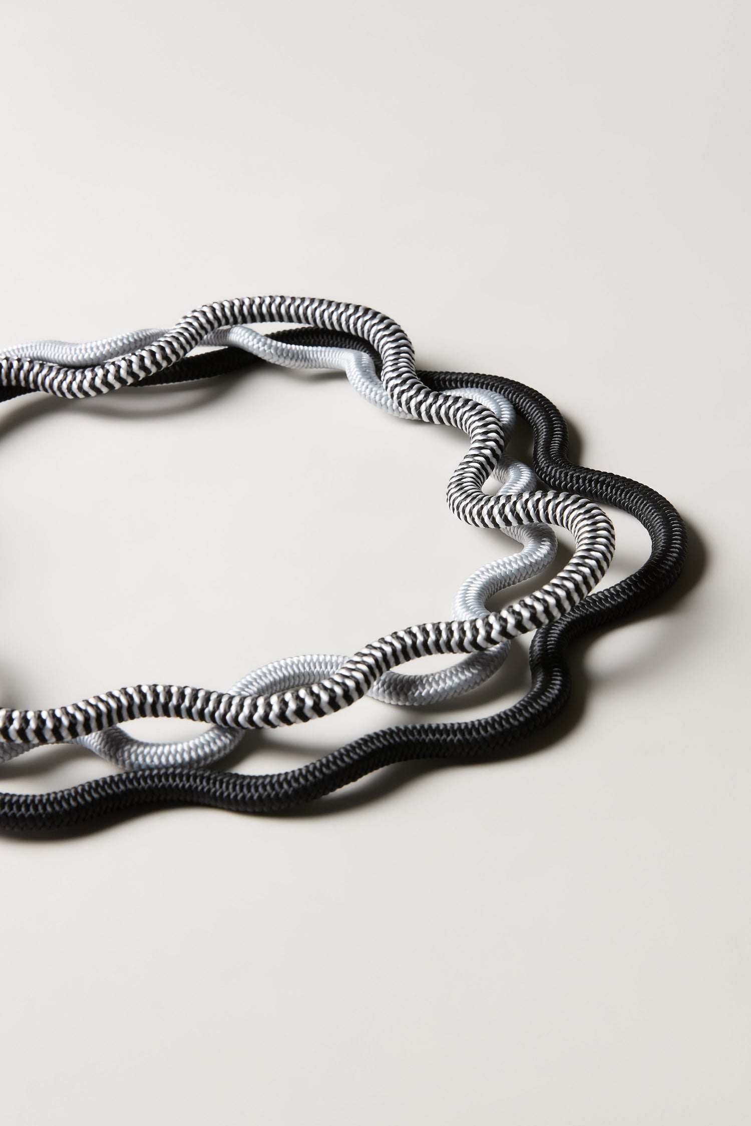 Two intertwined, wavy cords—one black with white patterns, one light gray—form an irregular oval on a light background, showcasing the sculptural style of the Black and White Monochrome Cord Waves Necklace.