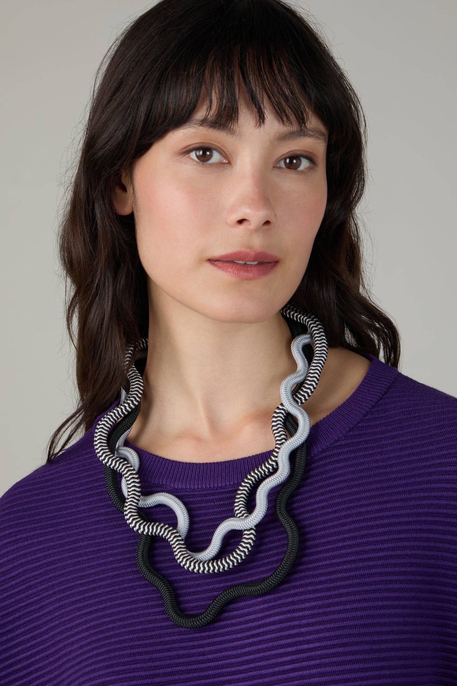 A woman with long dark hair, wearing a purple ribbed top and the Black and White Monochrome Cord Waves Necklace by Christina Brampti, looks at the camera.