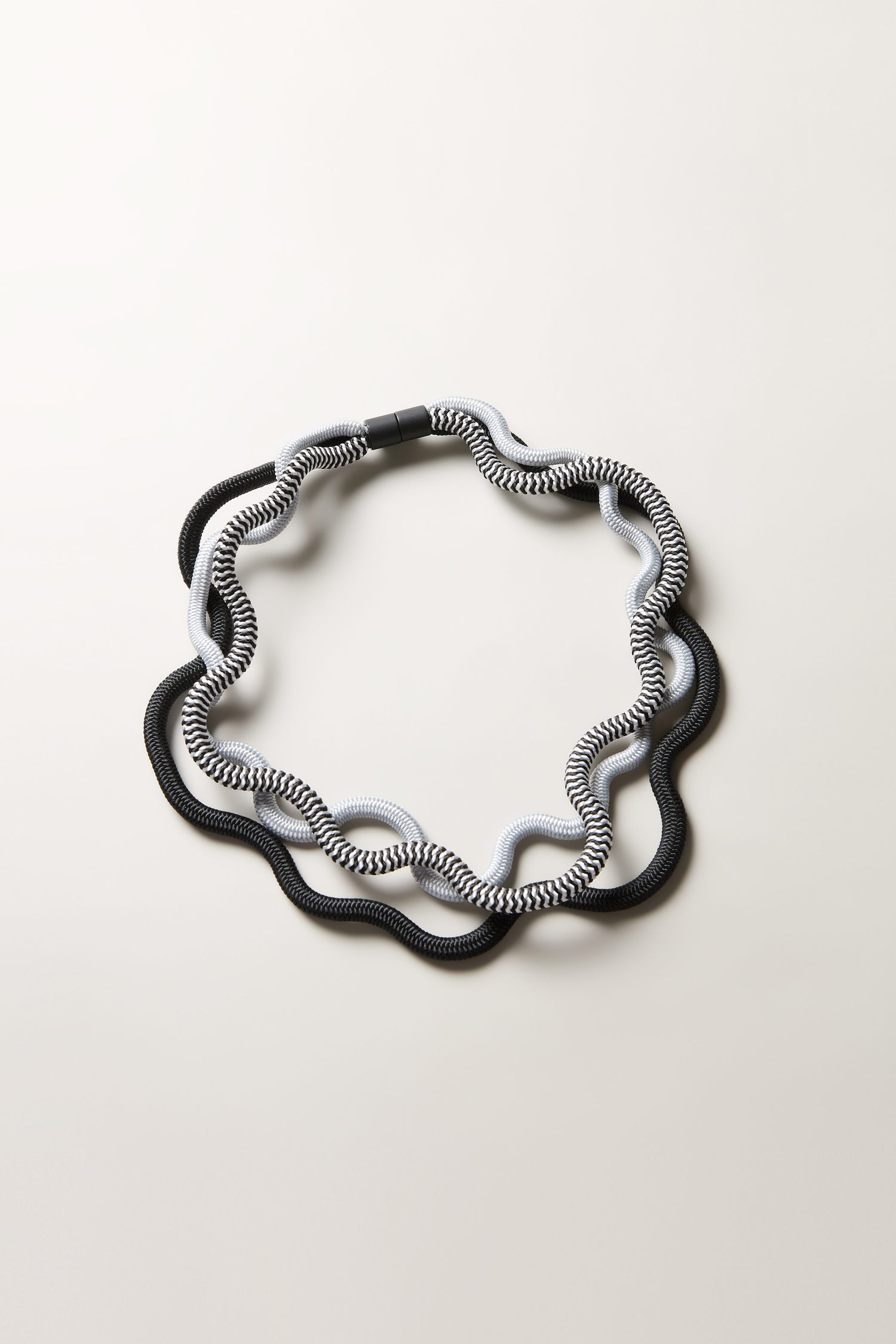 The Black and White Monochrome Cord Waves Necklace is laid flat on a light surface, its wavy circular form and alternating black, white, and gray segments resembling a sculptural jewelry piece or modern monochrome necklace.