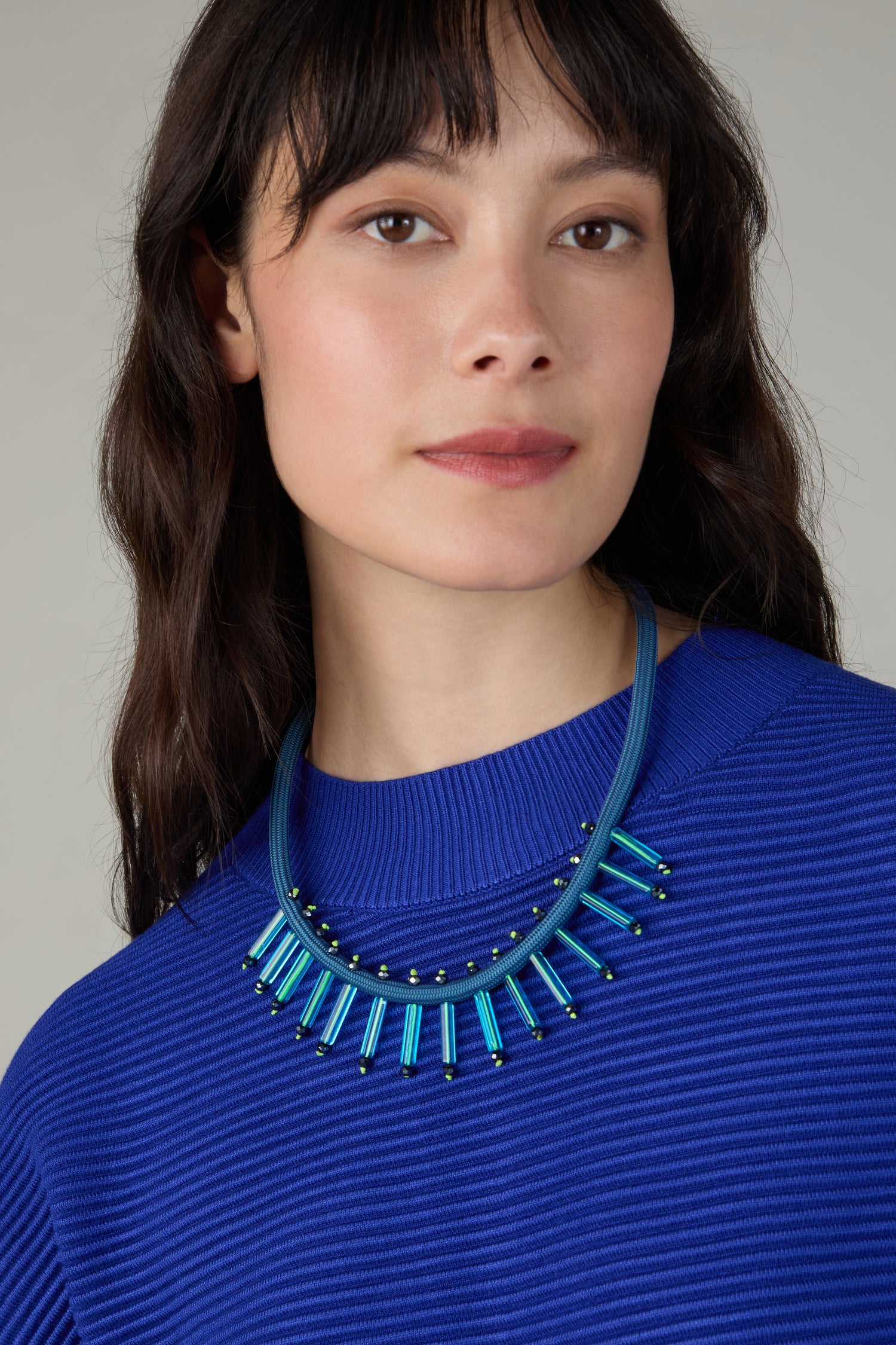 Woman with long dark hair wears a bright blue ribbed sweater and the Cosmic Necklace, a bold Christina Brampti design featuring contemporary blue and black jewellery elements, as she looks at the camera.