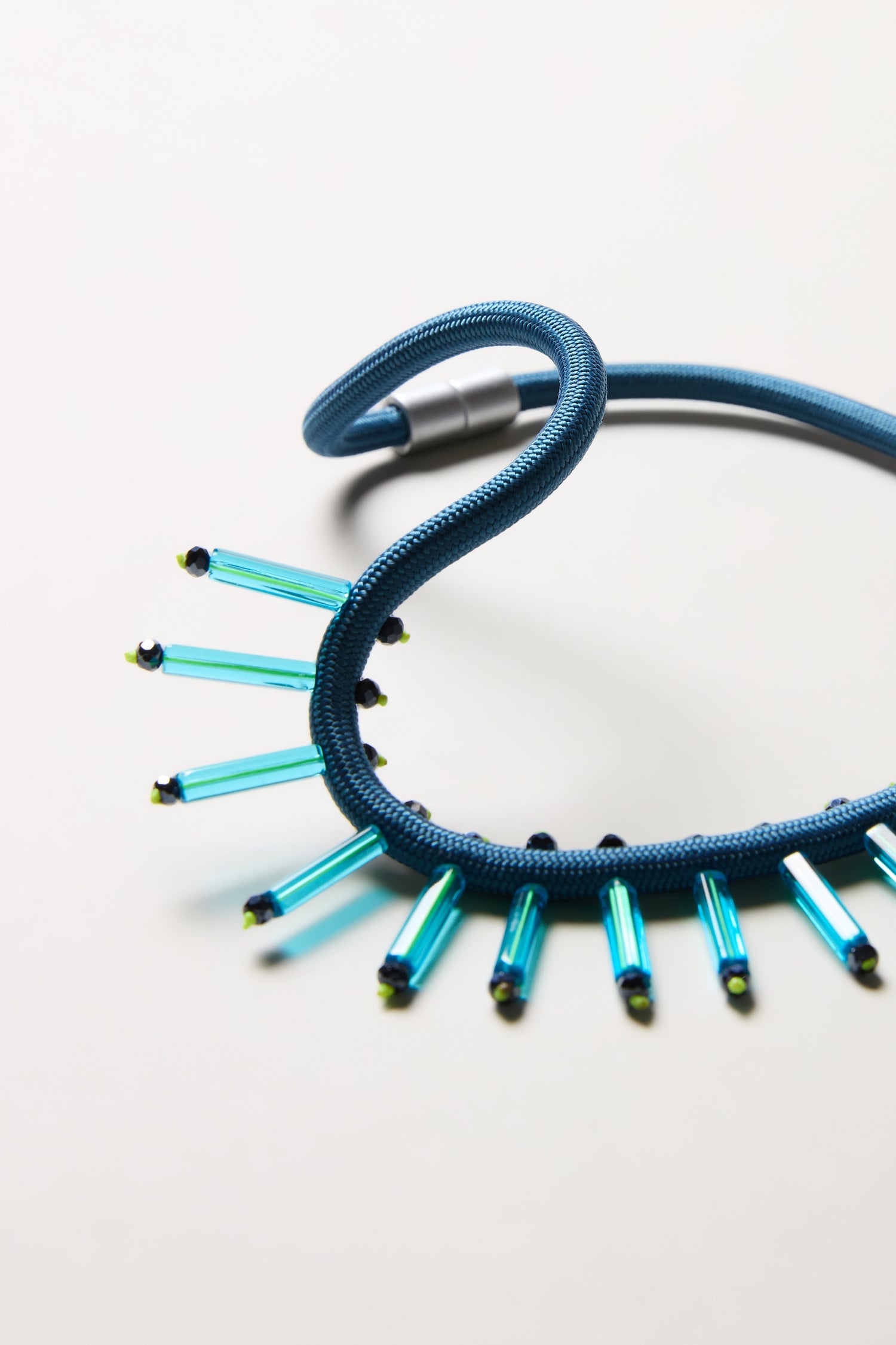 The Cosmic Necklace is a blue textured handcrafted piece featuring a metallic clasp and vertical turquoise rods with black and yellow ends along one side, displayed on a light surface—ideal for fans of contemporary jewellery.