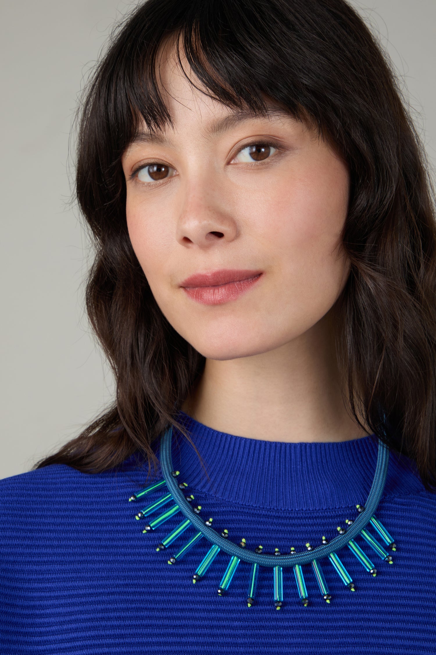 A woman with medium-length dark hair wears a bright blue ribbed top and the Cosmic Necklace—a bold blue piece with vertical tubular accents, reflecting her taste for contemporary jewellery.