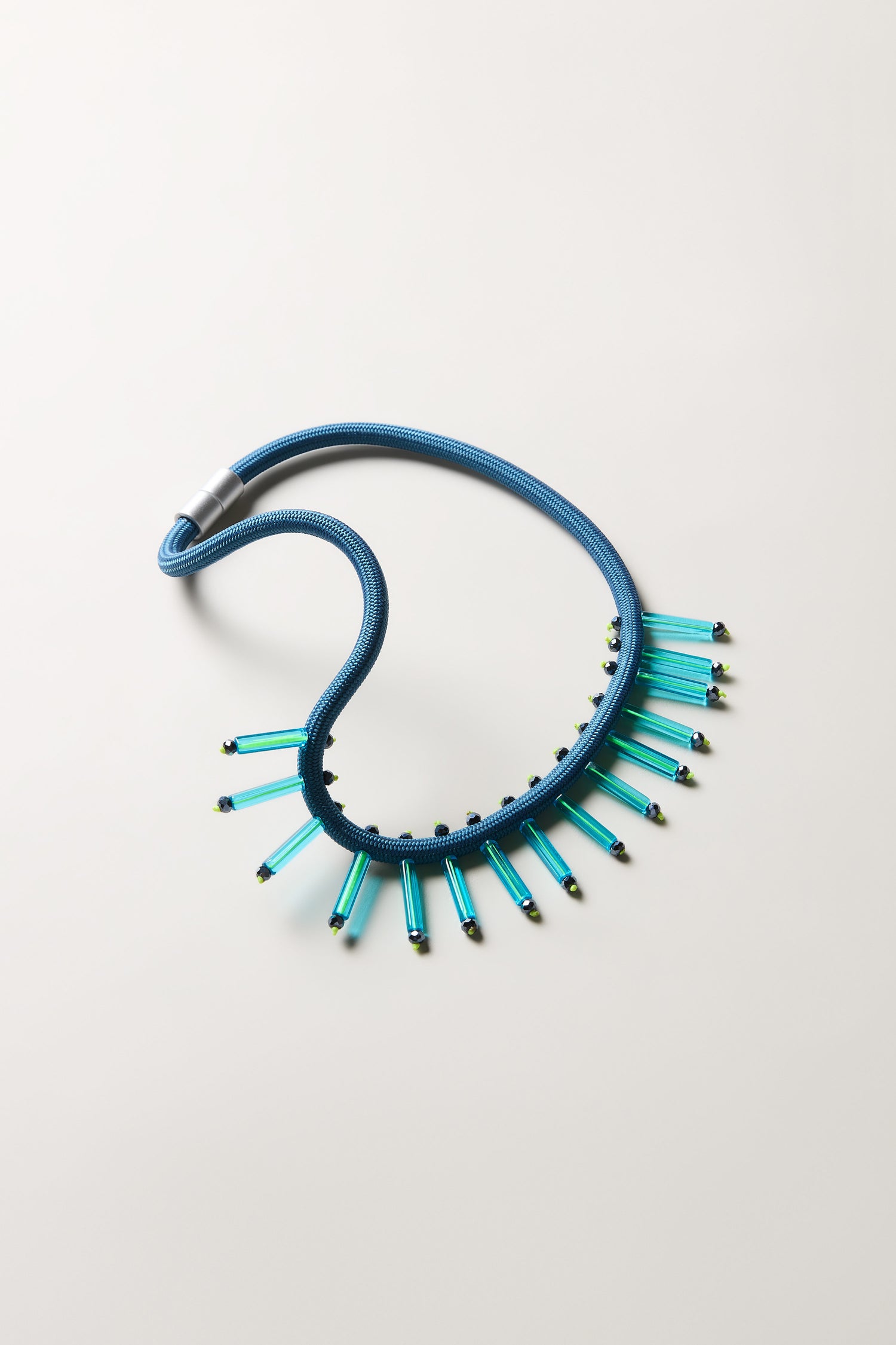 The Cosmic Necklace features a blue cord with evenly spaced, vertical blue-green cylindrical beads and black accents, handcrafted for a contemporary look and displayed on a light background—an eye-catching accessory.