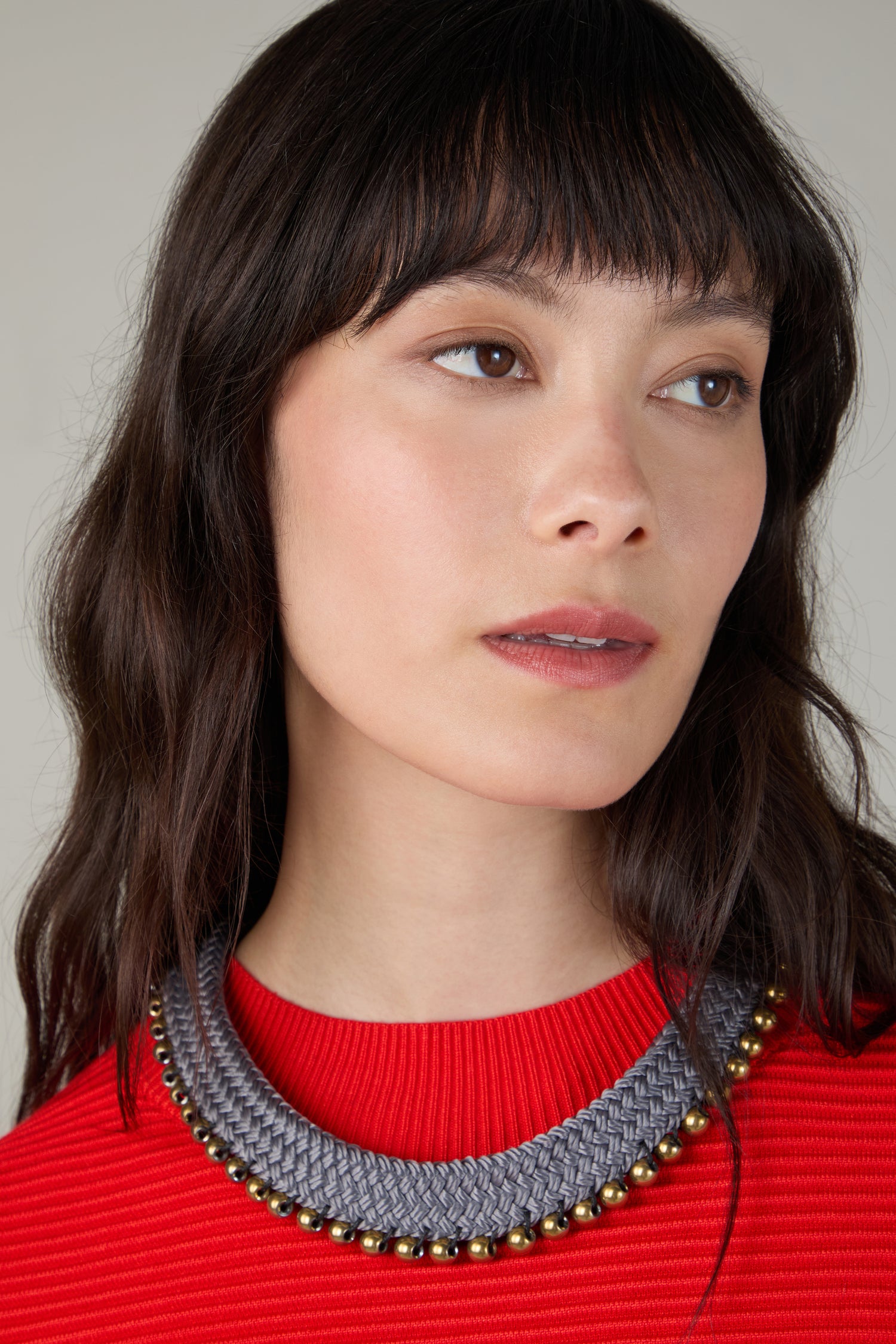 A woman with long dark hair and bangs wears a red ribbed top and a chunky Woven Soft Cotton Cord Necklace, looking slightly to the side.