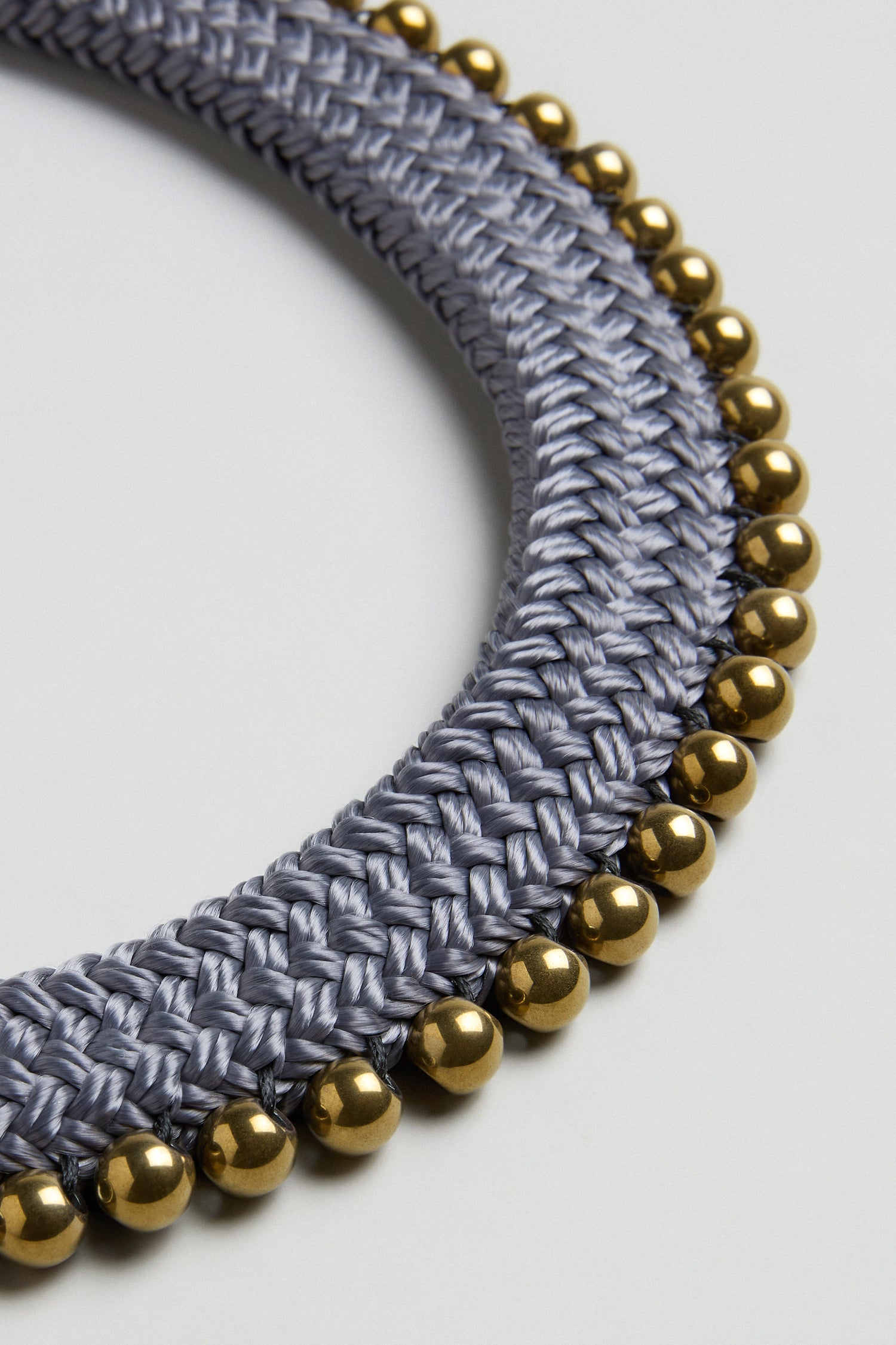 Close-up of the Woven Soft Cotton Cord Necklace by Christina Brampti, featuring braided silver-gray cords and a row of shiny gold beads along the outer edge, set against a plain background.