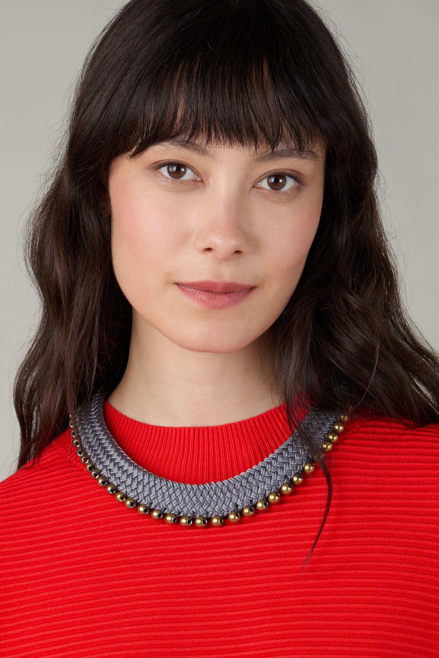 A woman with long dark hair and bangs poses against a neutral background, wearing a red textured top and a contemporary Woven Soft Cotton Cord Necklace with gold accents.