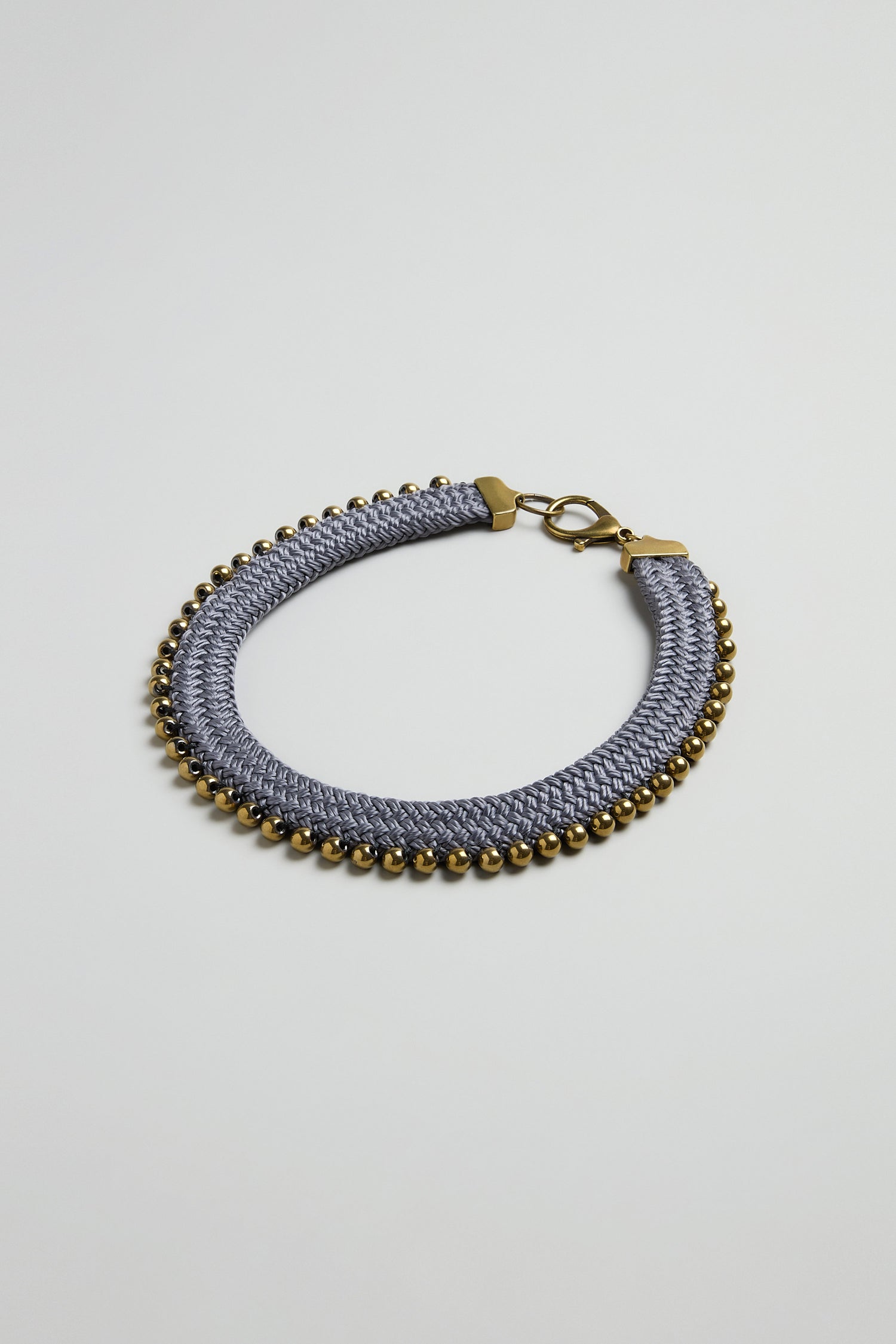 The Woven Soft Cotton Cord Necklace by Christina Brampti, in gray with small gold beads and a gold clasp, showcases contemporary jewelry style against a plain light background.