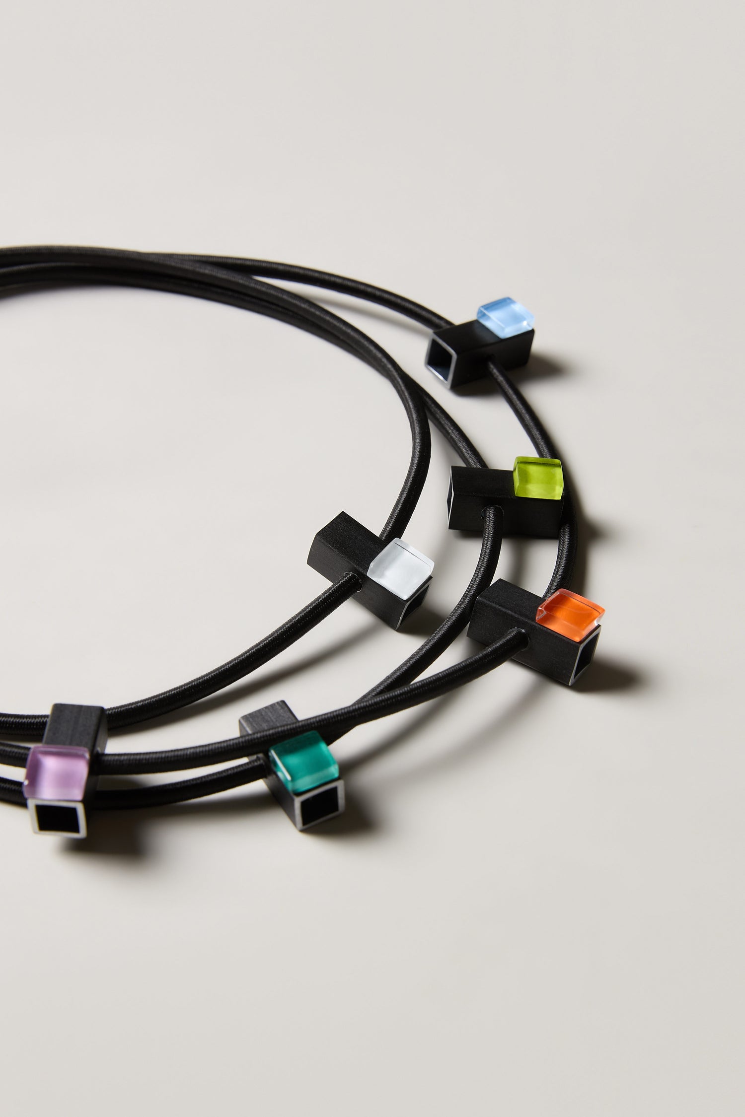 The Colour Pop Glass Beads Necklace features three black cords with cube-shaped pendants in blue, green, white, orange, and purple—modern statement jewellery inspired by Christina Brampti, shown on a neutral background.