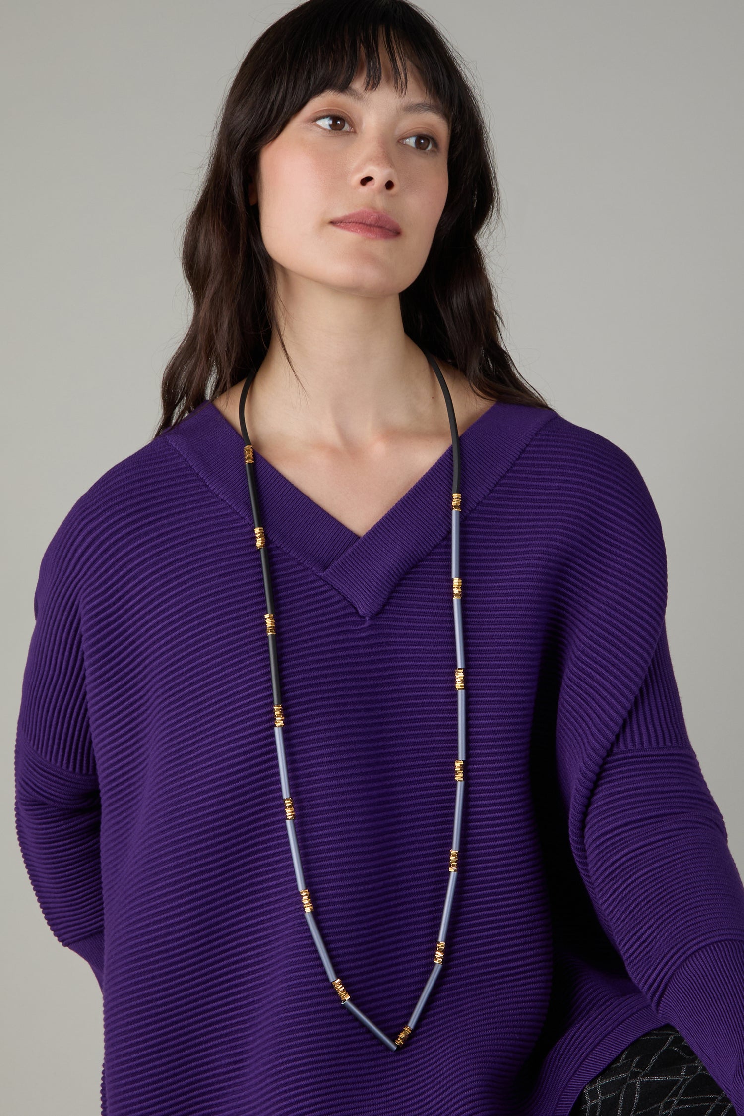 A woman in a ribbed purple sweater wears the Stacked Cylinders Necklace, a bold statement piece, and looks slightly upwards against a plain background.