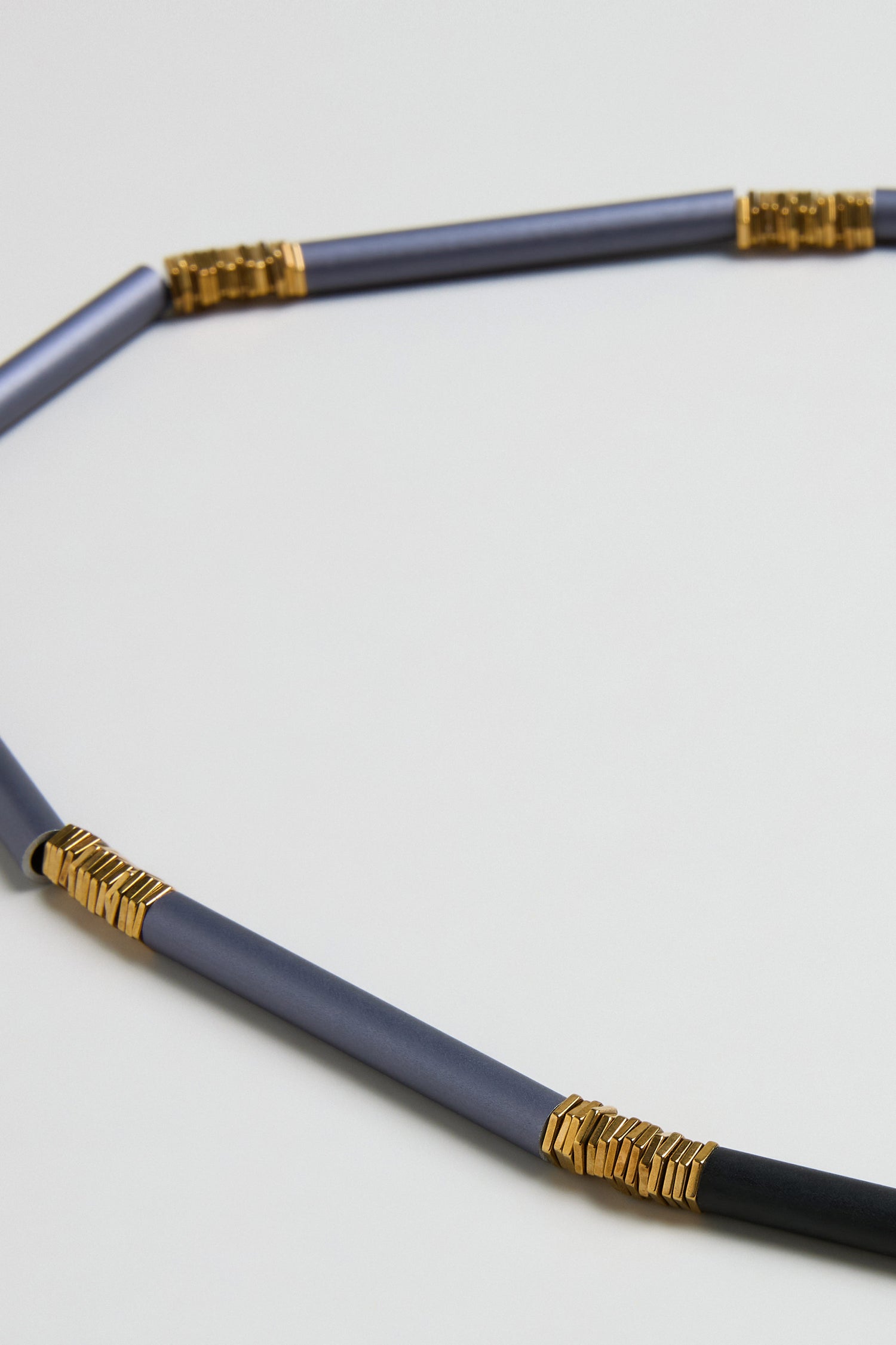 A close-up of the Stacked Cylinders Necklace, featuring metallic blue rods and gold-toned accents arranged in a contemporary partial circle on a light background.