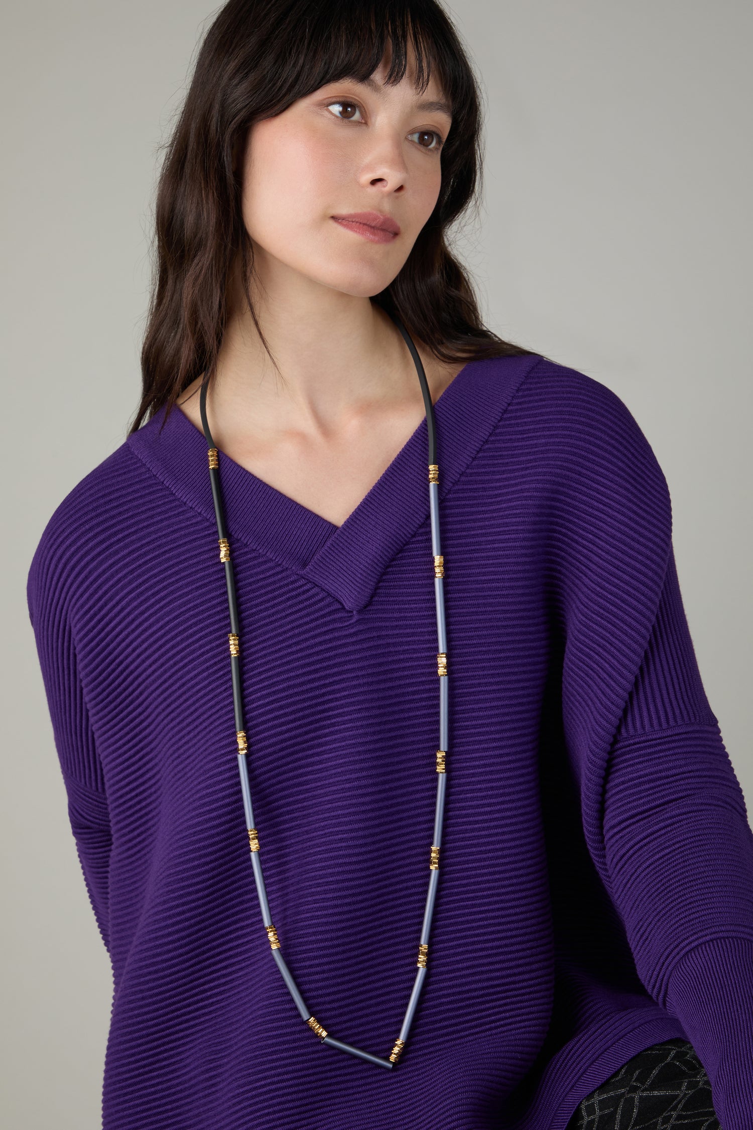 A woman in a loose-fitting purple ribbed sweater wears the Stacked Cylinders Necklace, looking slightly to the side against a plain background—a refined touch of contemporary jewellery.