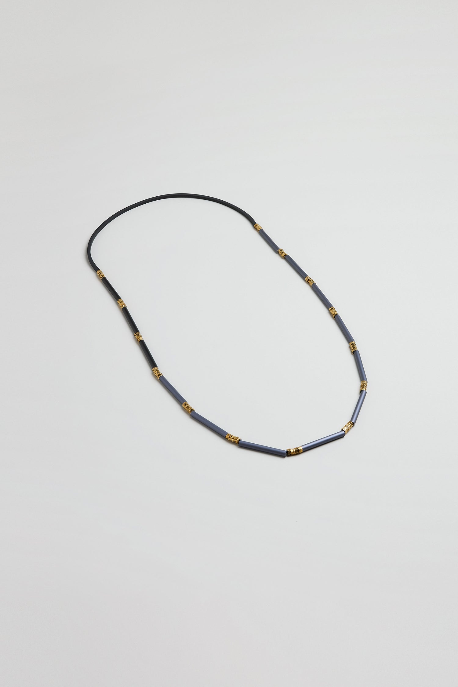 The Stacked Cylinders Necklace features alternating blue cylindrical beads and gold spacers on a thin black cord, embodying Christina Brampti’s modern style against a white background.