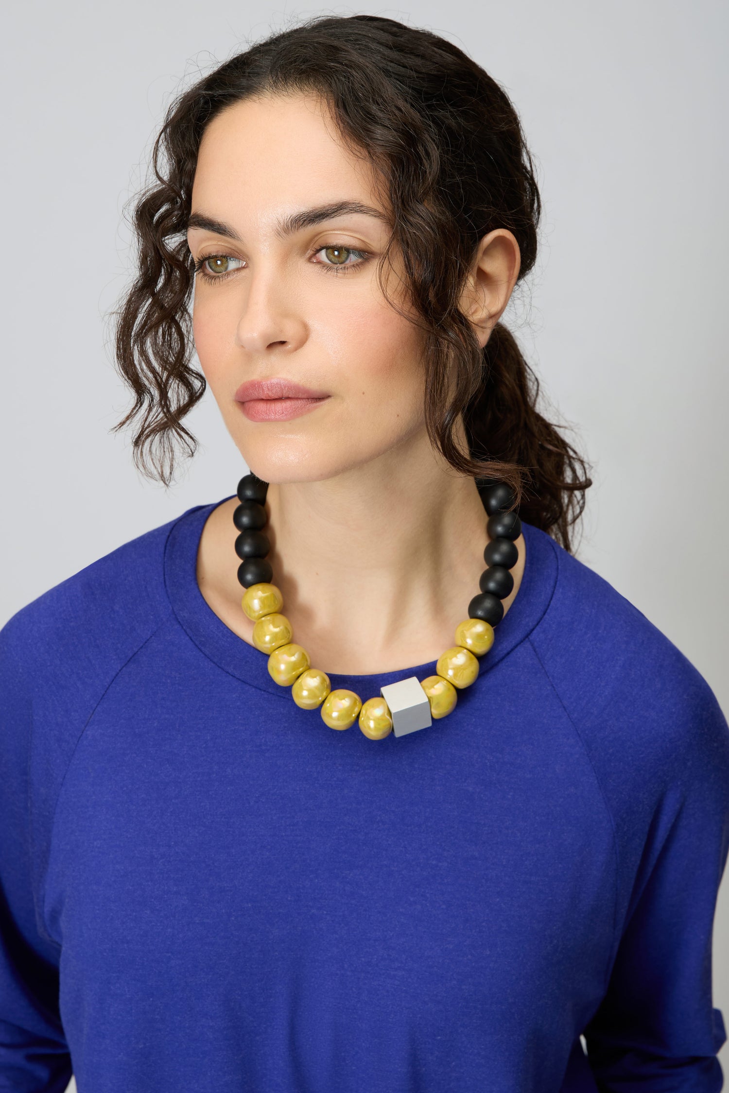 Woman in a blue top wears the Elements Necklace—a chunky, contemporary Christina Brampti piece featuring large yellow and black beads with a silver accent—while gazing slightly to the side against a plain background.