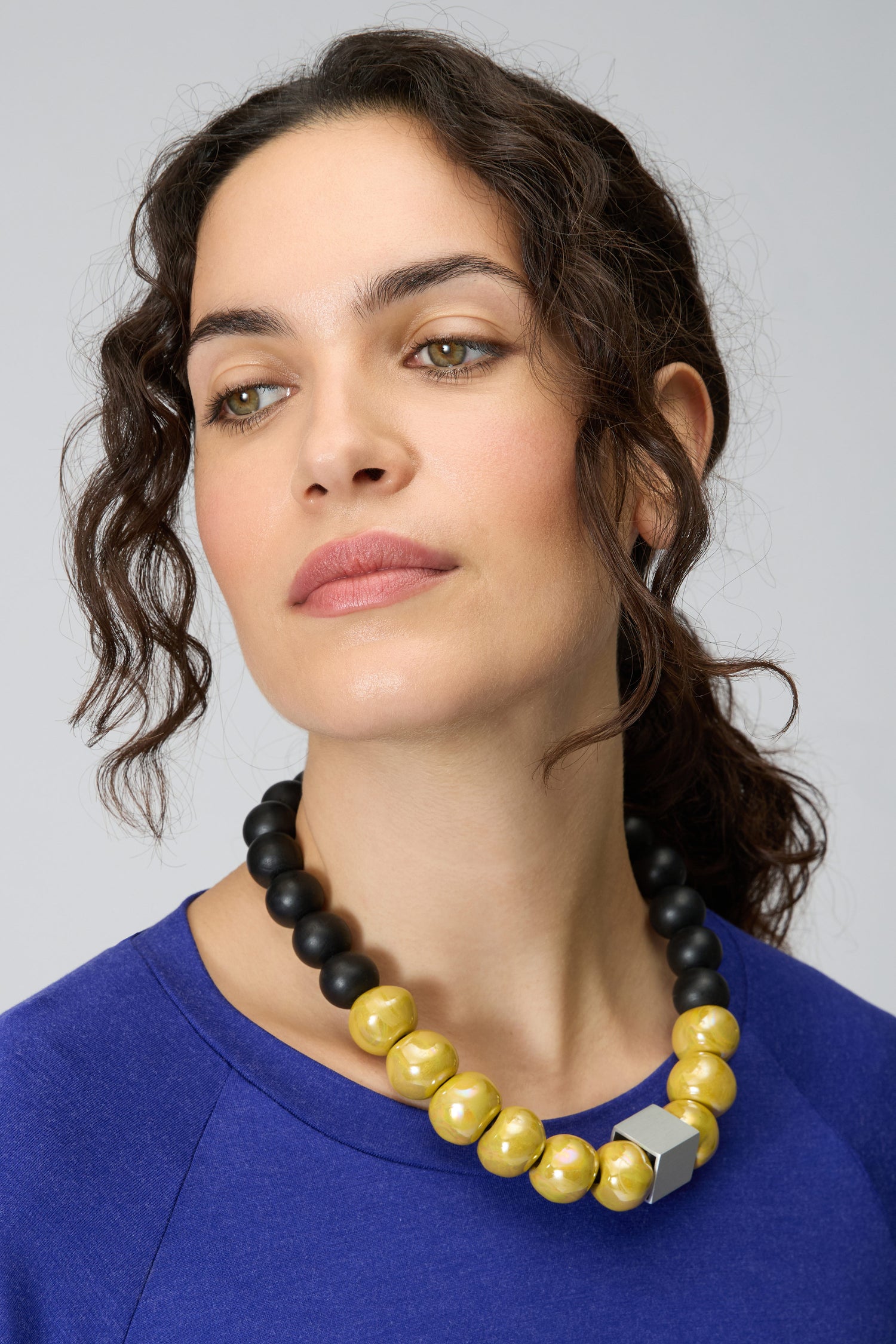 Woman with wavy dark hair in a blue top wears the Elements Necklace by Christina Brampti—an eye-catching piece of contemporary statement jewelry in yellow, black, and silver—while looking slightly to the side.