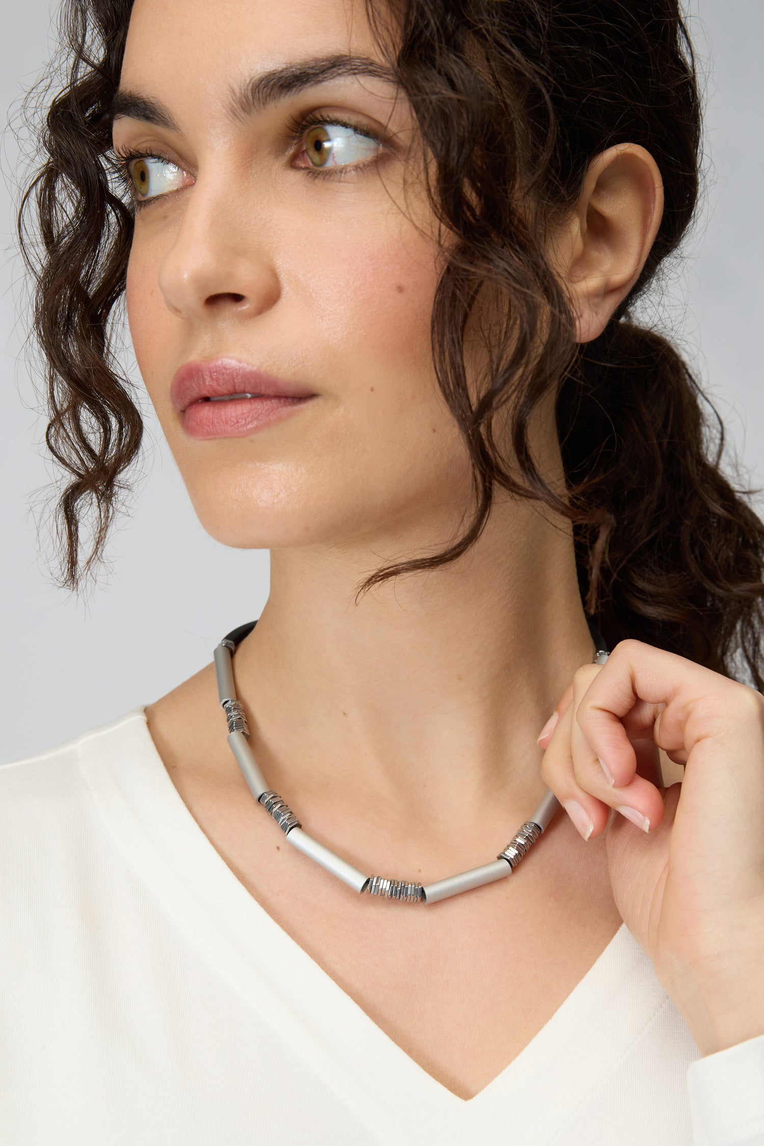 A woman with curly brown hair, wearing a white top, models the Silver Aluminium Hematite Necklace by Christina Brampti—crafted from silver, gray beads, and anodised aluminium—as she gazes to the side against a plain background.