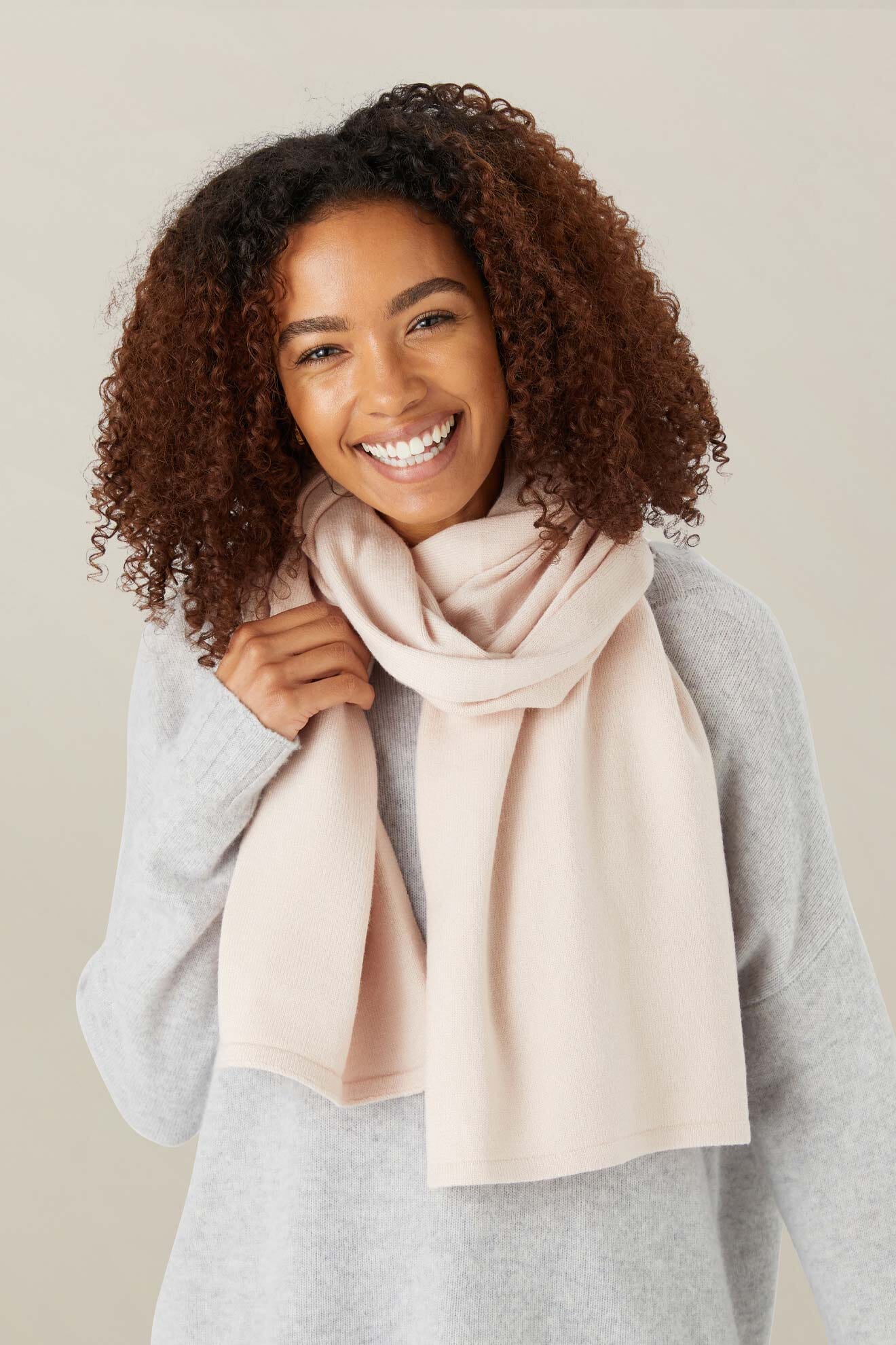 A woman with curly hair smiles at the camera, wearing a light pink Knit Scarf and a gray sweater against a plain background.